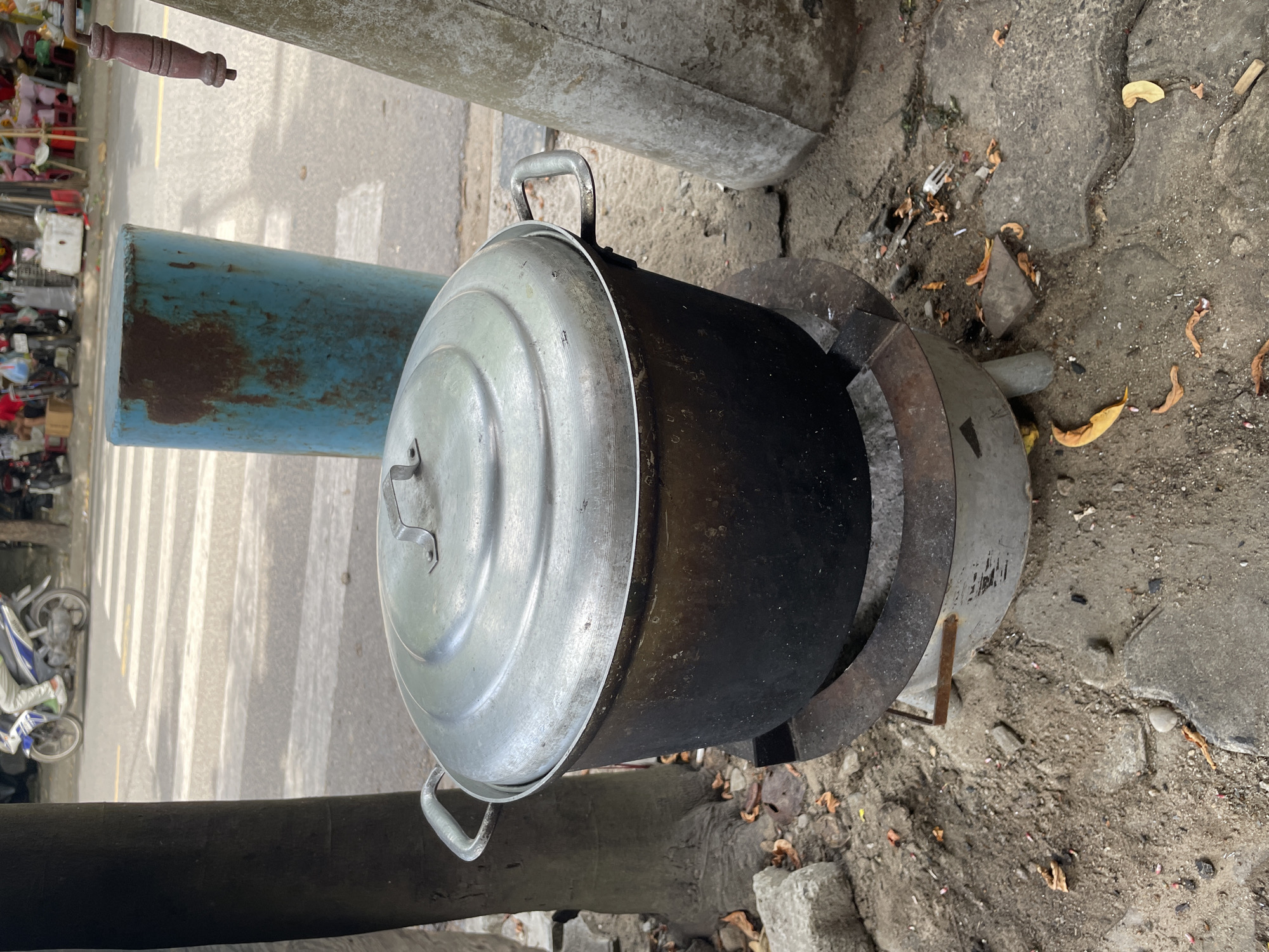 This image captures an outdoor scene in Đà Nẵng, Vietnam, likely on a street or sidewalk. In the foreground, a large, blackened metal pot with a shiny aluminum lid sits on a rustic metal stove. The pot shows signs of heavy use, with soot accumulated on its exterior. To the left, a section of a thick tree trunk is visible, and to the right, a textured, conical concrete structure is present. The ground is a mix of dirt, gravel, and concrete slabs, with fallen dry leaves scattered about. In the background, a pedestrian crossing with white stripes is visible on an asphalt road. Beyond the road, there are elements of a marketplace or street vendor area, with various items on display and motorcycles parked. A bright blue, cylindrical metal object, possibly a barrel, stands behind the pot. The overall impression is one of a functional, everyday outdoor cooking setup in a bustling urban environment.