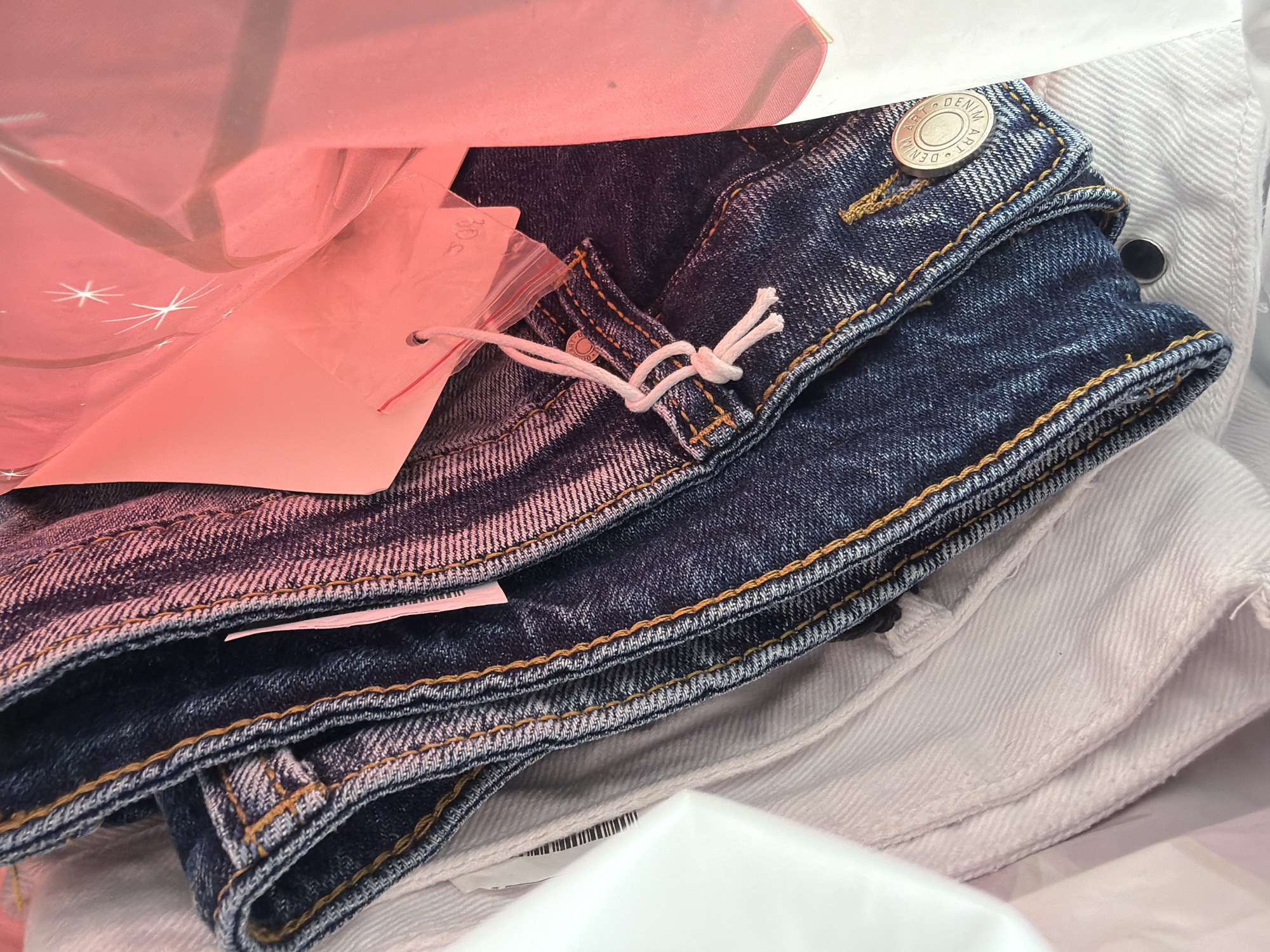 The image shows a pile of folded denim and white clothing items inside a shopping bag. A blue pair of denim jeans is on top, featuring a button closure with the text "ART DENIM" visible. A pink tag is attached to the jeans with a white string. The tag has a partially visible number "306" written on it. Underneath the jeans are folded white clothing items. The top of a pink bag with star patterns is also visible in the background. The location of the image is not discernible, nor any people, actions, or interactions in the scene. It cannot be verified that the location is in Shumen, Bulgaria.