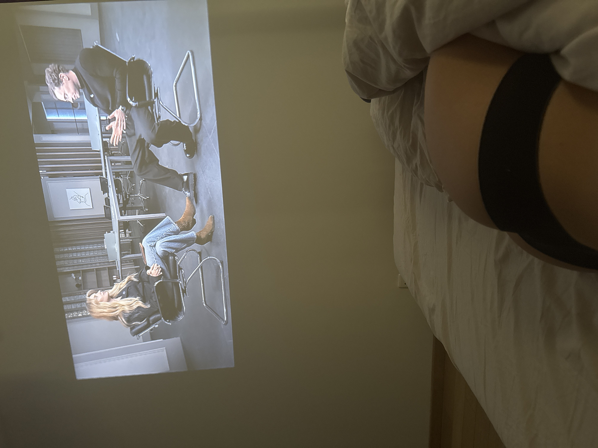 The image shows a person lying in bed under a white duvet, wearing black underwear. A scene from a television show or movie is projected on the wall adjacent to the bed. The projected image depicts two people sitting in chairs, seemingly engaged in a conversation or interview. The environment appears to be an interior setting, potentially a bedroom. There is no indication in the image that places it in Warsaw, Poland, or any other specific location.
