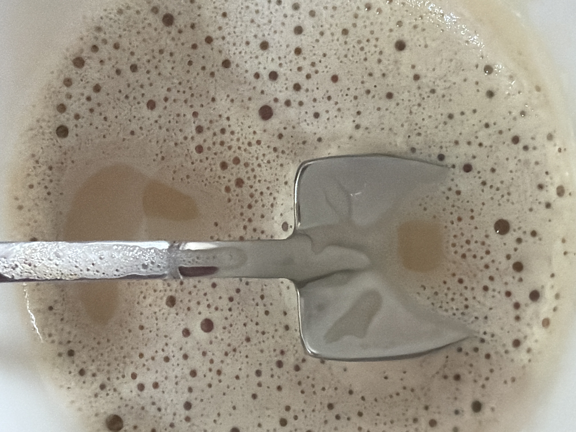 The image shows a close-up of a light brown, foamy beverage in a white container. A silver spoon rests inside the beverage, its head partially submerged. The surface of the drink is covered in small brown bubbles. No city or location is discernible.