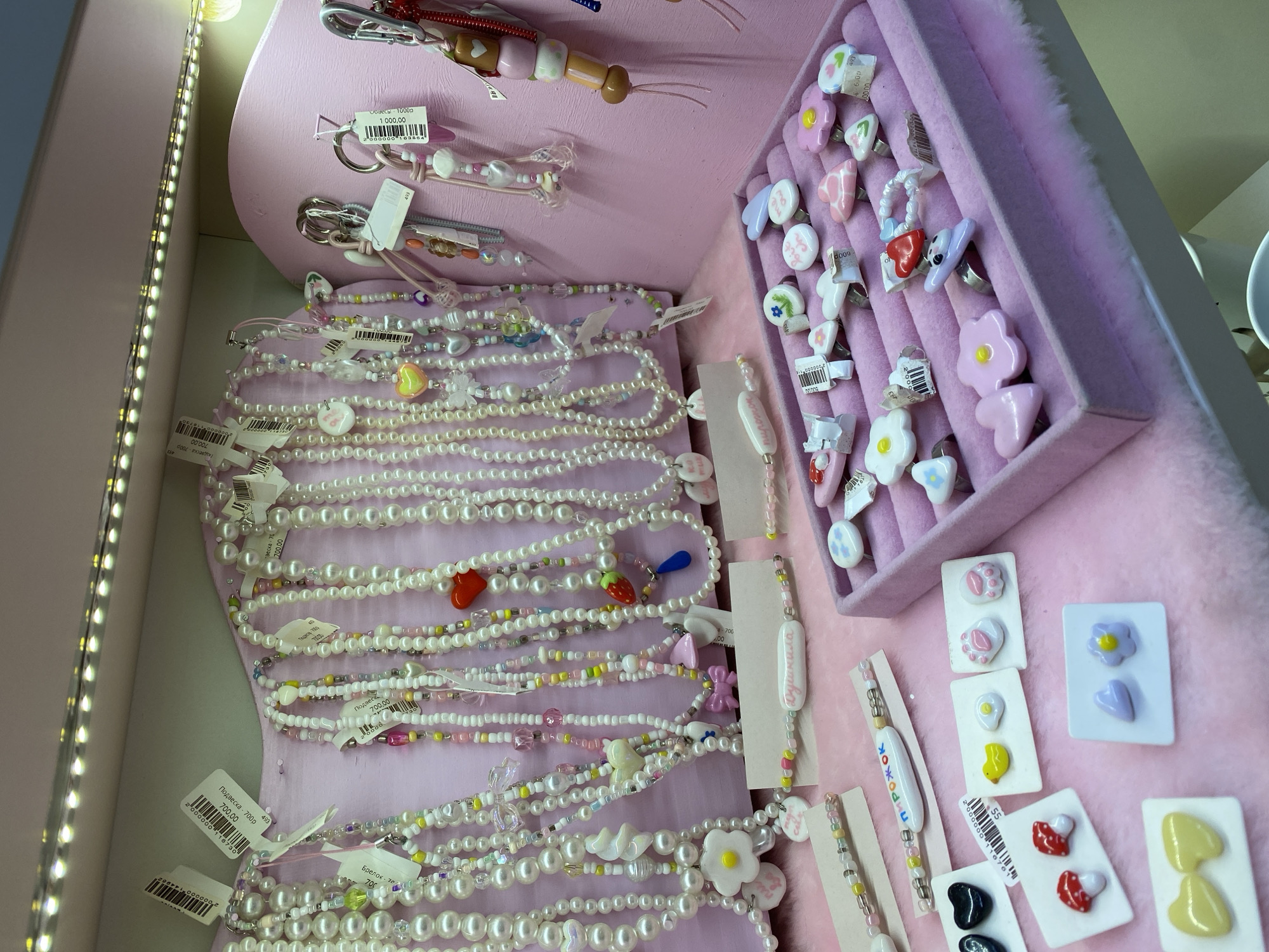 This image shows a display of jewelry, likely for sale, within a store or market setting. The dominant color scheme is pink, with a pink backdrop and display cases lined with pink fur-like material. The jewelry includes pearl necklaces with colorful beads and charms, rings with flower and animal designs, and earring sets mounted on small white cards. Price tags in Russian are visible on many items, suggesting a location in Russia or a Russian-speaking area. The items are arranged in an organized manner within the display, with the necklaces cascading on the left and the rings and earrings presented in trays and on cards to the right. A strip of LED lights illuminates the scene from the side, enhancing the visibility of the jewelry. The overall impression is a vibrant and potentially artisanal collection of accessories. The exact location in the city of Unknown, Unknown is not knowable from the image.