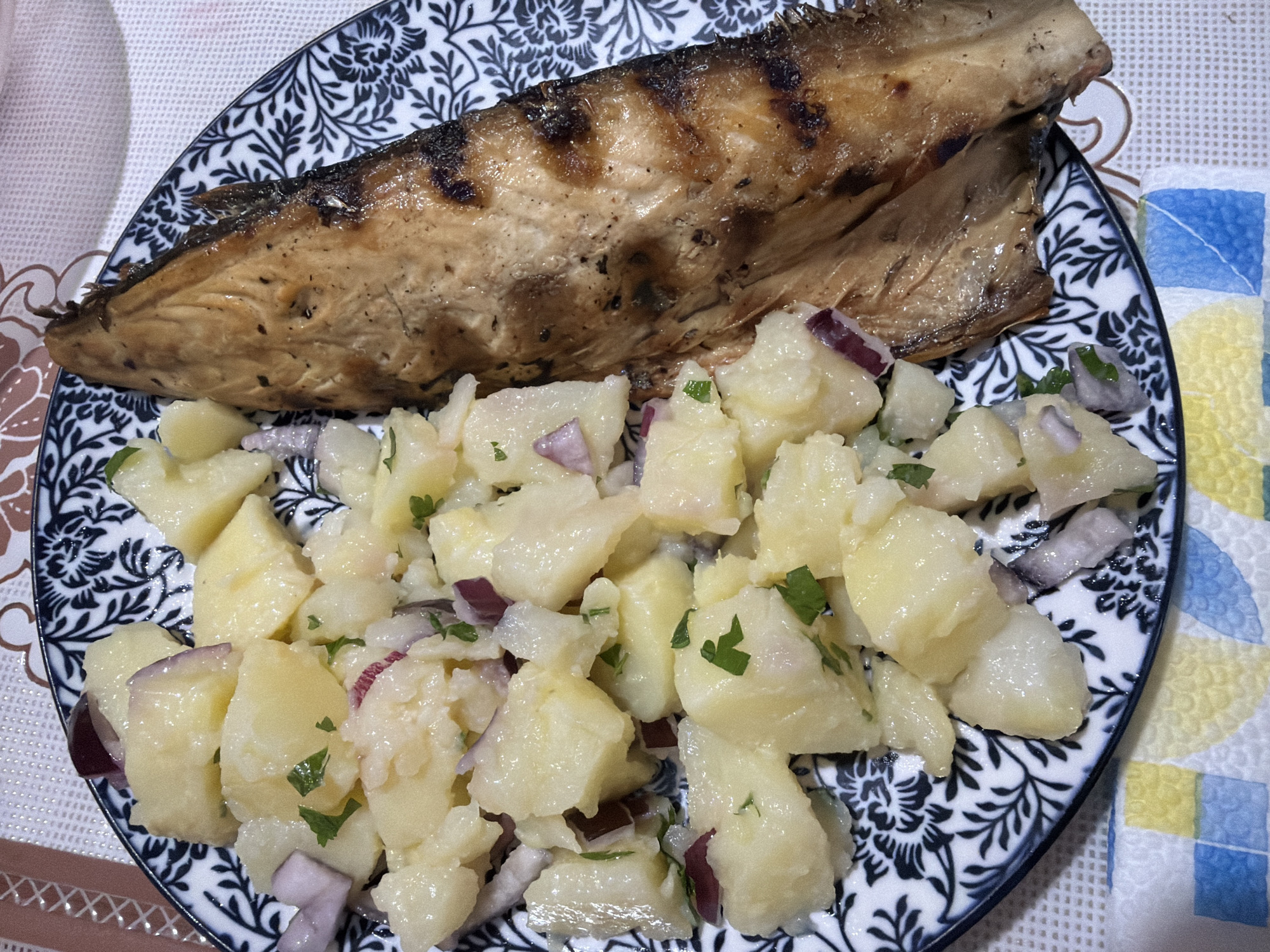 A close-up, overhead shot shows a serving of potato salad and a piece of grilled fish on a patterned plate. The potato salad is composed of cubed potatoes, chopped red onion, and parsley, all dressed in a light vinaigrette. The grilled fish, likely mackerel based on its shape and coloration, is skin-on and shows distinct grill marks. The plate has a black and white floral pattern. The plate rests on a placemat with a textured white surface and a colorful geometric pattern visible at the bottom. The scene is likely a meal served in a home kitchen or a restaurant in Pazardzhik, Bulgaria, though no specific location within the city is identifiable from the image alone.