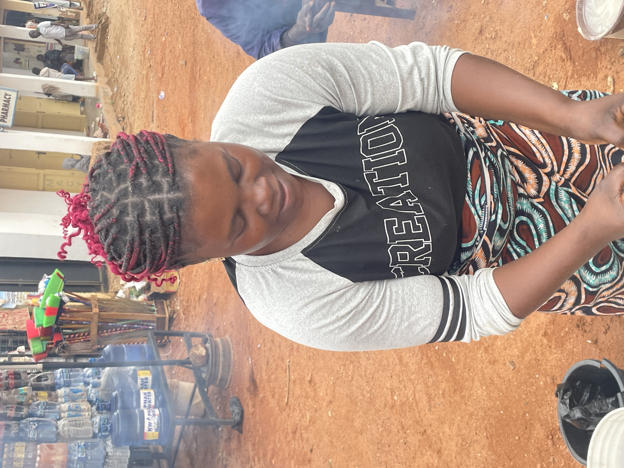 A medium-shot photograph shows a woman sitting outdoors, likely in Jos, Nigeria. The woman has dark skin, braided hair with red accents, and is wearing a grey and black baseball tee with "RECREATION" printed on the front. She is wearing a patterned skirt.
The woman appears to be preparing food or working with her hands, as indicated by the presence of cookware and steam. To her left is a rack filled with bottled water and cleaning supplies. In the background is a pharmacy.
The ground is a reddish-brown dirt. Other people are in the background.