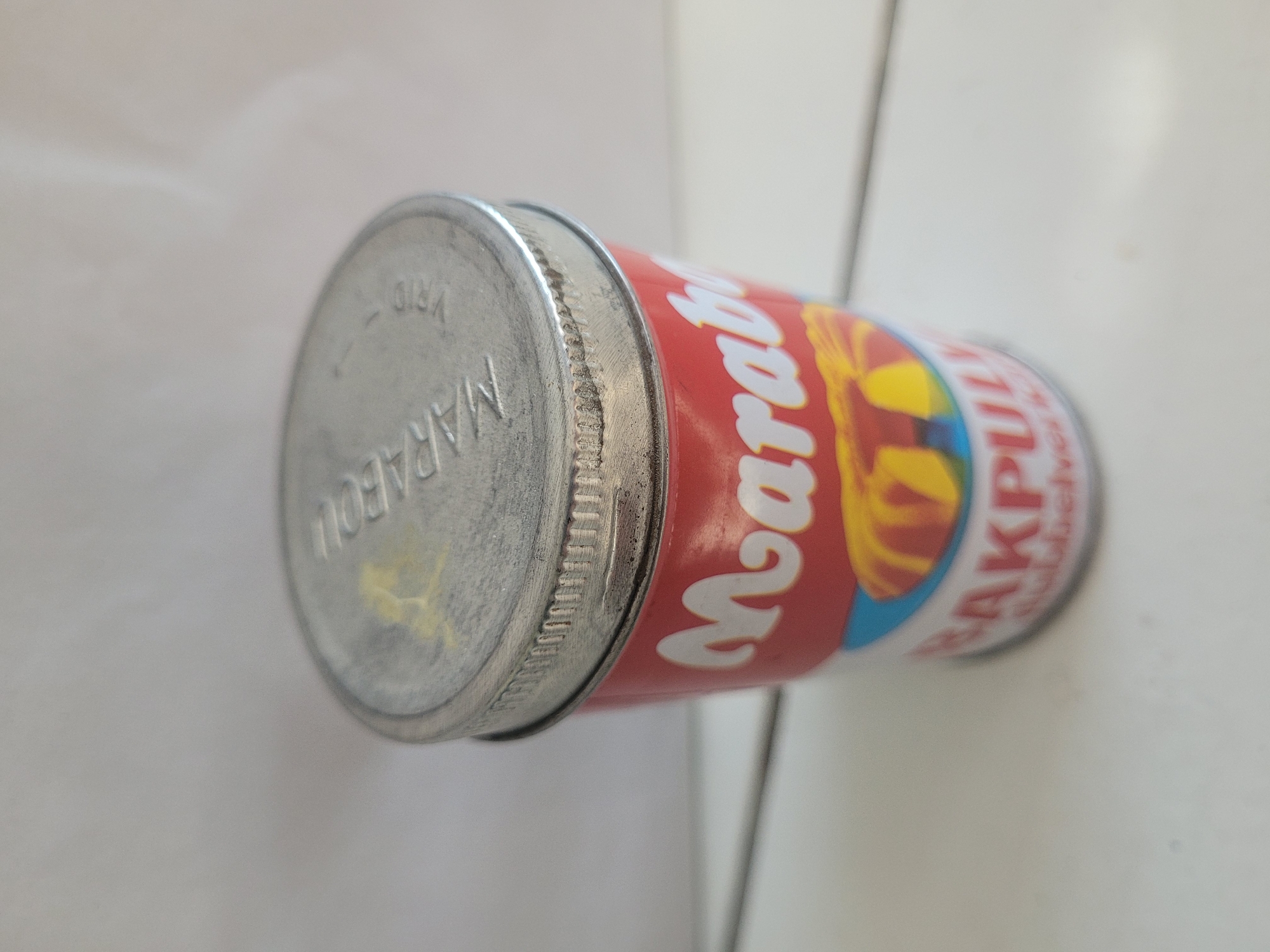 A vintage tin of Marabou baking powder, labeled "MARABOU BAKPULVER", rests on a white, wooden surface. The tin is predominantly red with white lettering for "Marabou" and yellow and blue accents depicting what appears to be a cake or dessert. The silver lid has embossed lettering that is partially obscured. The background is a plain, light-colored wall. The scene is indoors and the lighting is diffuse. This image does not contain any information that would allow for the identification of its location within the city of Gävle, Sweden.