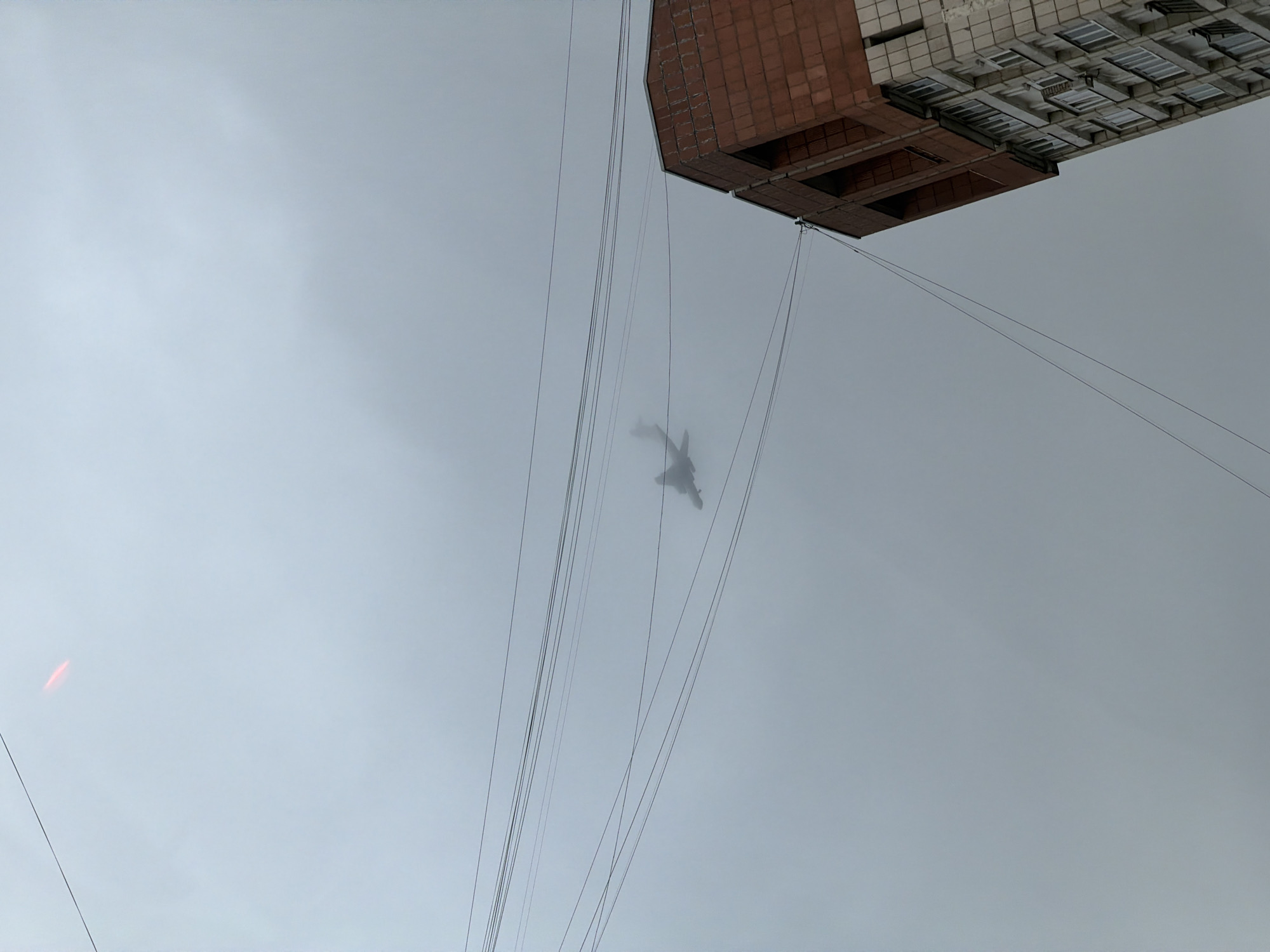 The image presents an upward view of an urban landscape under a heavily overcast and hazy sky. In the upper portion, a multi-story building is partially visible. It features a facade primarily constructed from light-colored square tiles or bricks, with a distinct, darker reddish-brown section below. Multiple rectangular windows are discernible across the building's surface.
Numerous thin, dark cables or power lines stretch downwards from the vicinity of the building, diagonally crossing the frame against the pale sky. Amidst this network of wires and partially obscured by the thick atmospheric haze, a shadowy, human-like figure is suspended. The figure appears to be in a somewhat crouched or seated position, possibly attached to the cables, and is quite indistinct due to the low visibility.
The sky is uniformly light gray, indicative of a very cloudy or foggy day, which creates a soft, diffused light and contributes to the overall mysterious atmosphere. A small, elongated, blurry red light or object is faintly visible in the lower-left corner of the frame. The scene suggests a potential activity involving aerial work, a stunt, or an art installation within an urban environment. There is no visible text in the image. Given the context, the location is Perm, Russia.