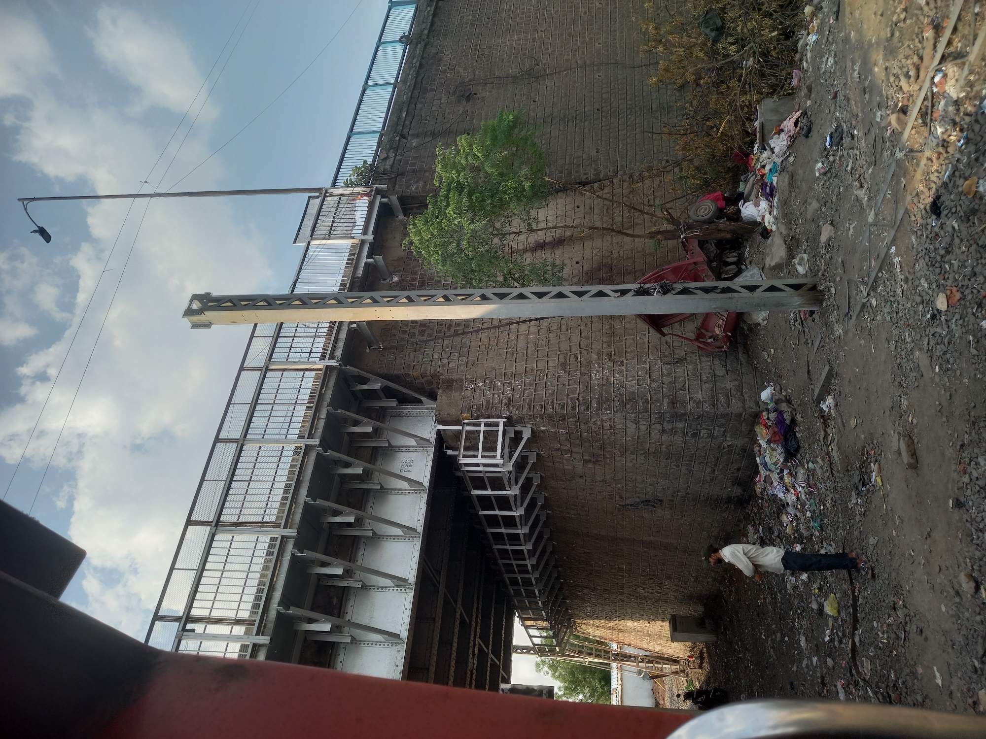 The image depicts a scene in Bhopal, India, dominated by an elevated structure, likely a railway overpass or flyover. The structure is constructed of stone and steel, with a stairway attached to its side. Debris and discarded items are scattered on the ground next to a person lying down. A streetlight and power lines are visible above. A tree grows out of the wall. A red vehicle is overturned.