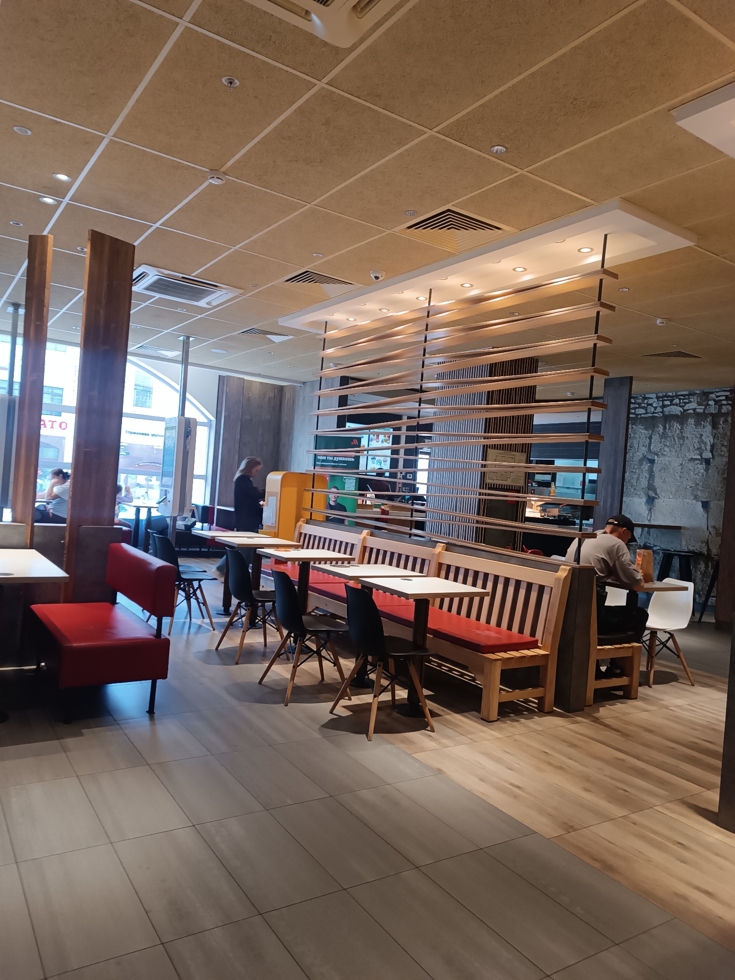 The image shows the interior of a McDonald's restaurant in Tula, Russia. The restaurant has a modern design with wooden accents and bright lighting. There are several tables and booths, some of which are occupied by customers. In the foreground, a red booth is visible, and further back, a man in a black cap is sitting at a table. The ceiling is made of acoustic tiles and features recessed lighting and air vents. The walls are a mix of concrete and wood paneling. A large window overlooks an outdoor area.