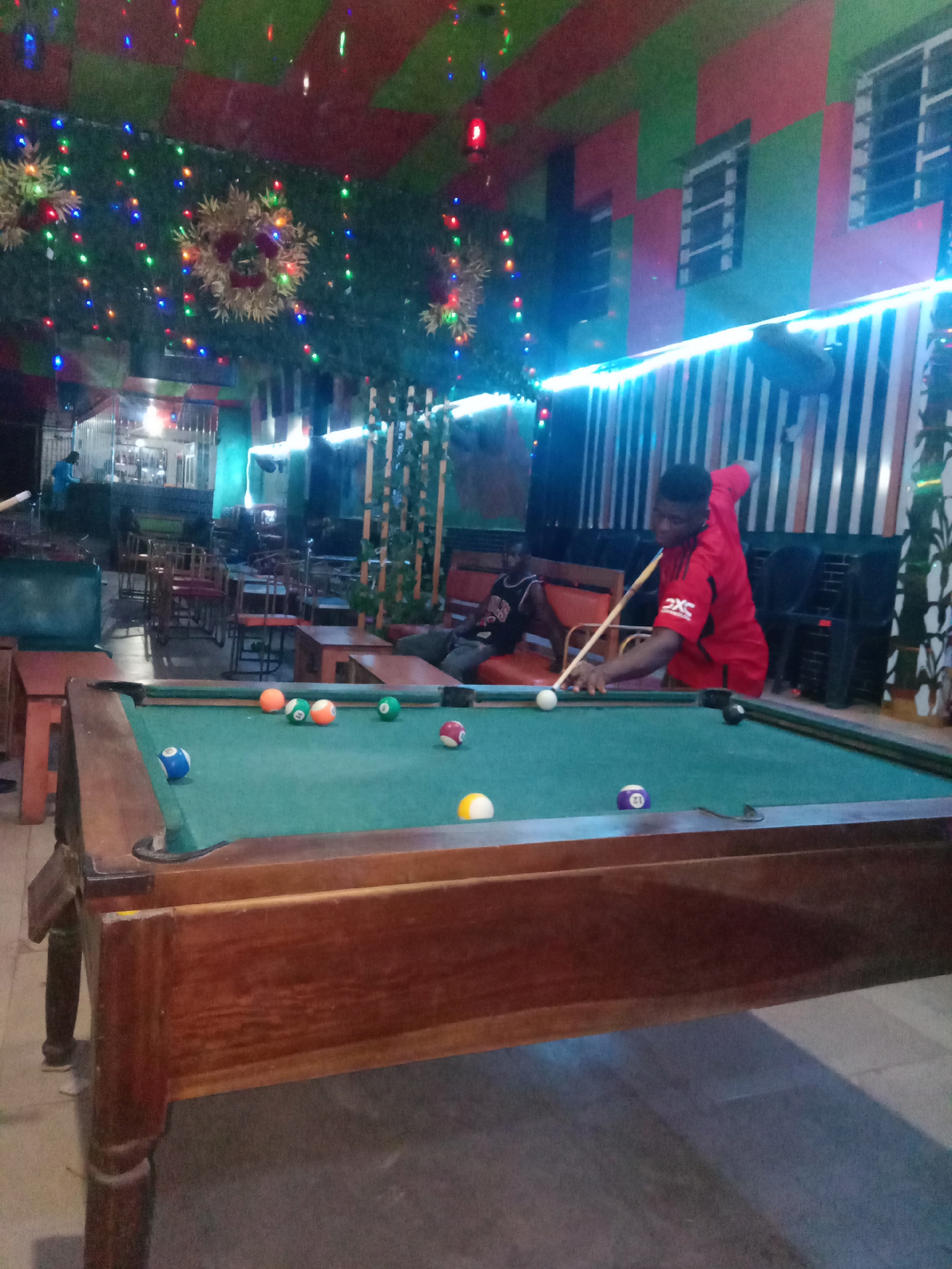 A photograph captures an indoor scene in what appears to be an entertainment venue in Onitsha, Nigeria. A man in a red shirt is positioned at a pool table with several billiard balls scattered across its green surface. He is in the act of taking a shot, cue stick in hand. Another individual is seated in the background. Decorations are visible in the surroundings, including lights and floral arrangements hanging from the ceiling, adding a festive atmosphere to the room. The walls are patterned with a combination of green and red hues. Furniture, including chairs and tables, is scattered throughout the space.