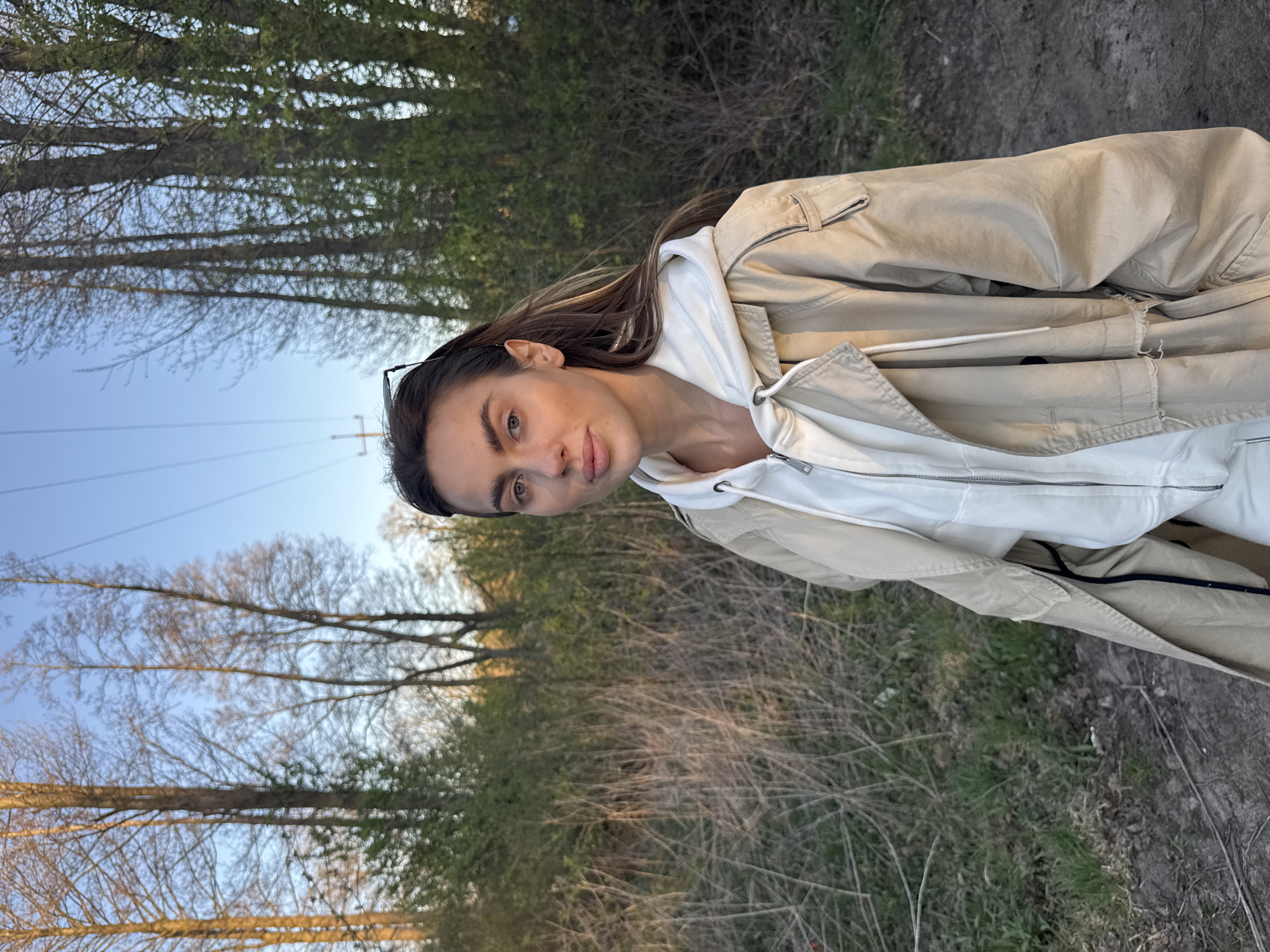 A woman stands outside on a dirt path with grass and some shrubbery. A line of trees is along the left side of the image and a blue sky can be seen above the trees. The woman is wearing a white hoodie and a tan jacket. She is facing the camera with her head tilted slightly. The image appears to have been taken at Arciechów, Poland.