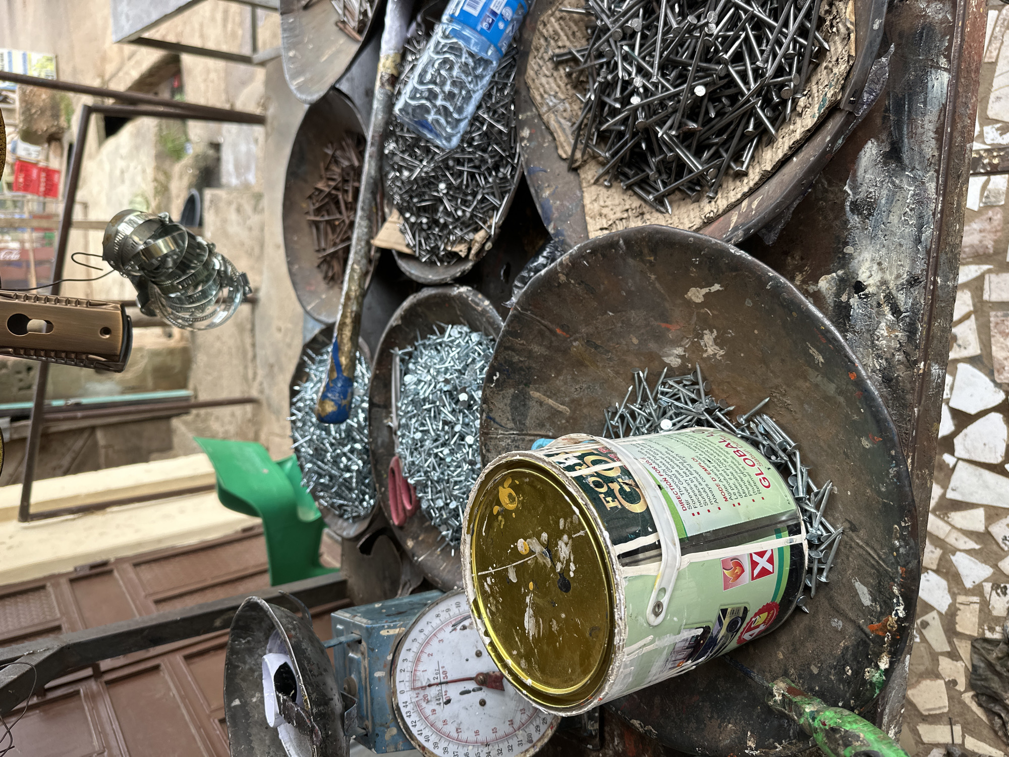 This image captures a close-up view of a market stall in Narayi, Nigeria, filled with various hardware supplies, primarily nails and other metal fasteners. In the foreground, a large, partially used can of "Forever Gold Gloss" paint sits on a worn, metal surface. The can's label, partially obscured, indicates it is a paint product with instructions in English and French. Around the paint can and in several metal bowls, a substantial quantity of assorted nails is displayed. Some bowls are filled with silver-colored nails, while another contains darker, possibly rustier, nails. A vintage scale, blue and white, with a circular dial showing measurements in pounds and kilograms, is positioned to the left of the paint can. Behind these items, other market elements are visible, including a green plastic chair, a metal object hanging from above, and in the background, a doorway or entrance to a building with a tiled floor. The overall scene suggests a busy outdoor or semi-enclosed marketplace where construction or repair materials are sold.