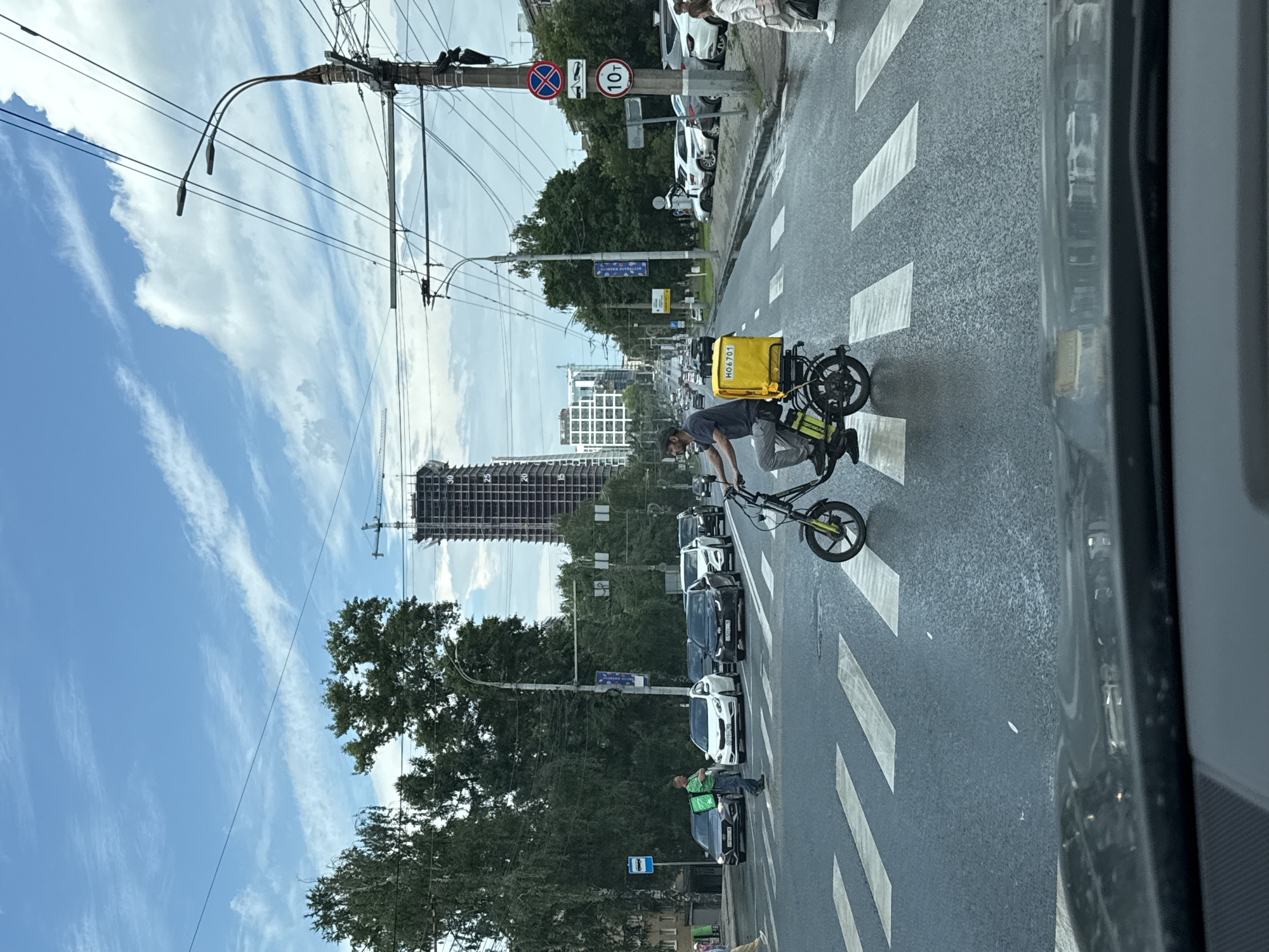 The image depicts a street scene in Izhevsk, Russia, under a cloudy sky with hints of blue. In the foreground, a person on a yellow electric scooter with a yellow delivery box is crossing a pedestrian crosswalk. The scooter appears to be a three-wheeled model. The person is wearing dark clothing and a cap.
The setting is an urban environment with a road, several lanes of traffic, and a pedestrian crossing. Cars are stopped at the crosswalk, waiting for the scooter rider to pass. In the background, there is a tall, unfinished building with exposed structural elements, suggesting ongoing construction. Trees line the street, and utility poles with numerous wires are visible.
Signage in the image includes traffic signs, one of which indicates a 10-ton weight limit. Another sign appears to be a blue bus stop sign. A yellow sign on the delivery box displays the text "HO 6701".
The overall impression is a snapshot of everyday urban life, possibly during the daytime, with a delivery person navigating traffic. The lighting suggests it's likely not early morning or late evening.