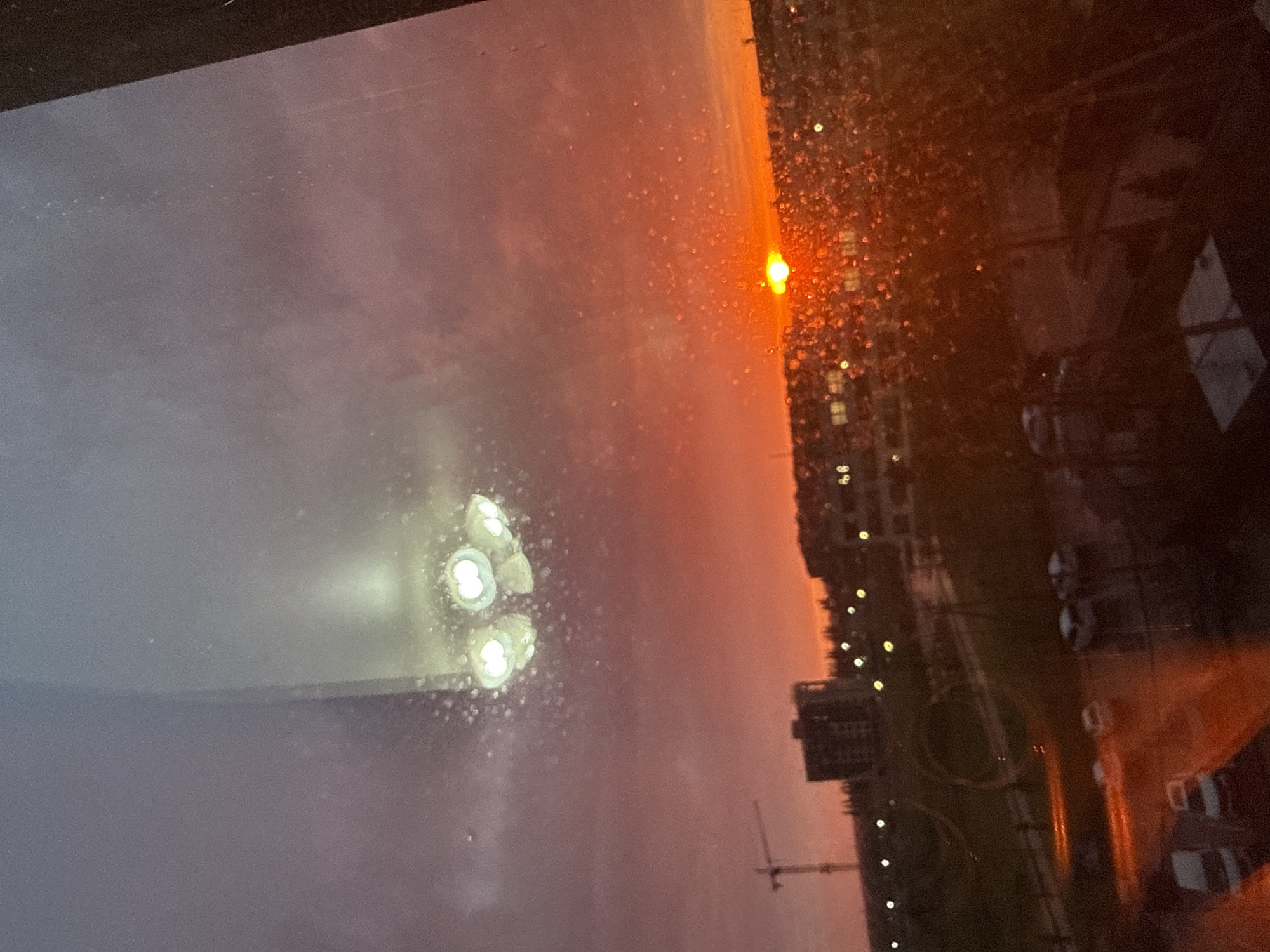 The image shows a dusk scene in an urban environment, viewed through a rain-splattered window. The sky is a mix of orange, red, and purple hues. Outside the window, buildings and a crane are visible. Reflections of interior lights are also seen on the window.