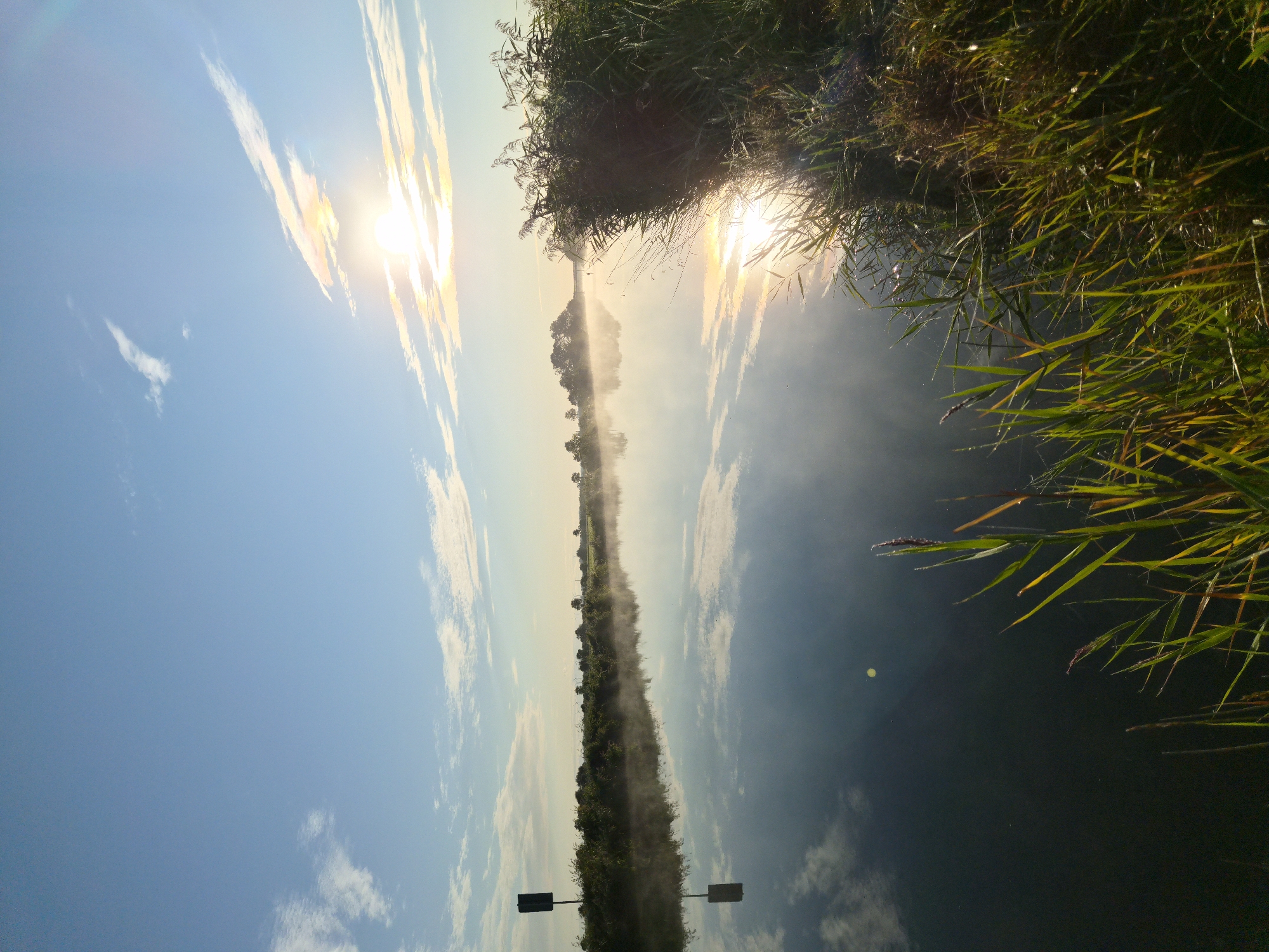 This is a vertical, eye-level, outdoor photograph of a calm river on a sunny day. The sun is positioned in the upper right quadrant, partially obscured by wispy clouds, casting a bright glare on the water. The sky is a clear blue with scattered white clouds. A narrow strip of land with green vegetation lines the left bank of the river, featuring a dark, rectangular marker post. On the right bank, lush green reeds and tall grasses grow densely. The water reflects the sky, clouds, and the distant treeline with a slight mist rising from its surface. The overall atmosphere is serene and peaceful. The location is the Geeste river in Schiffdorf, Germany.