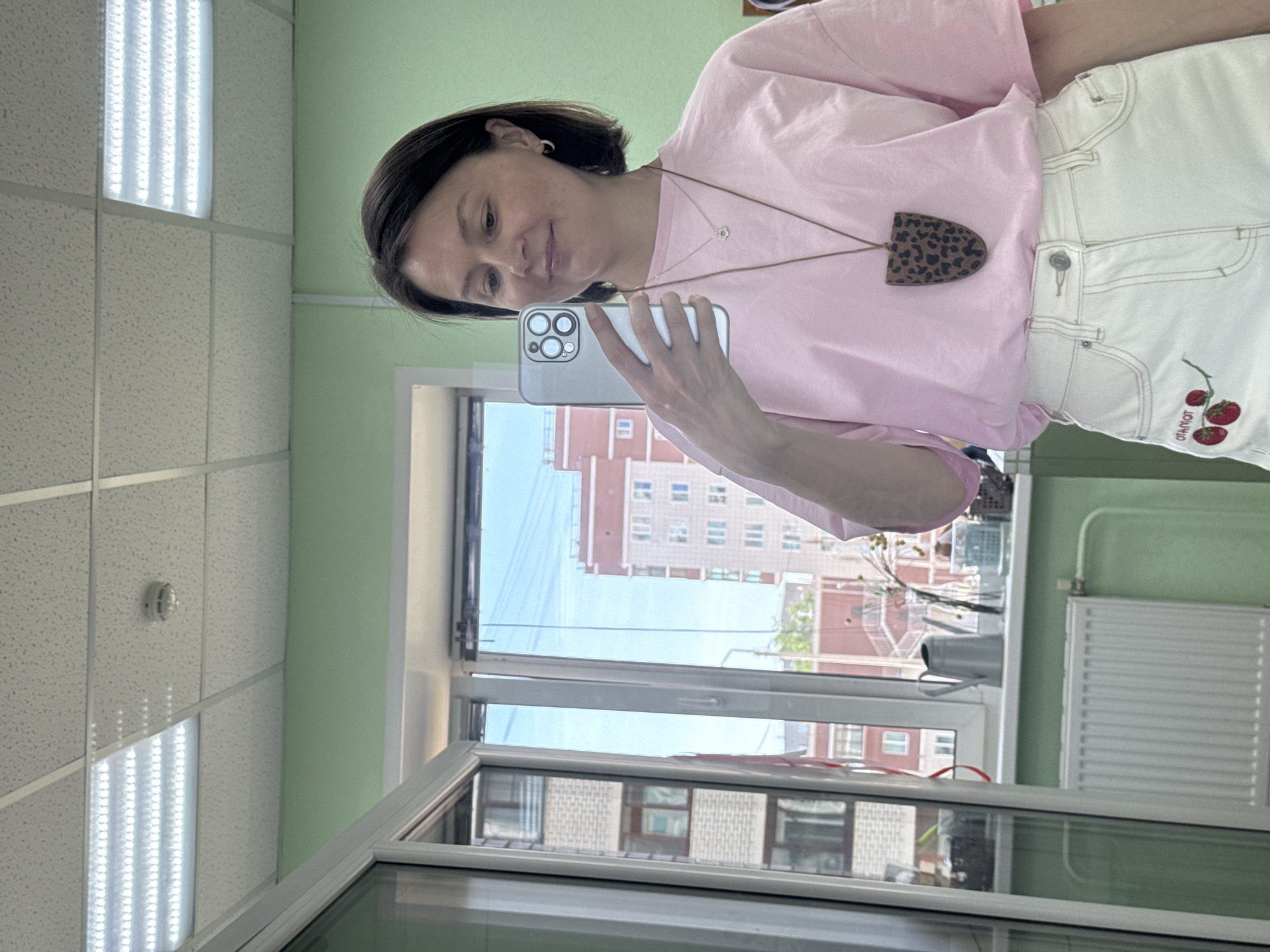 A woman is taking a selfie in a room with a window. The room has a drop ceiling with fluorescent lights, and green walls. Outside the window is an urban scene. From what is visible, the architecture of the buildings in the background suggests this is Izhevsk, Russia. The woman is wearing a pink t-shirt and white pants with red strawberry embroidery. She is holding a smartphone in her left hand to take the photograph. She also has a necklace with an animal print pendant. A radiator is against the right wall beside the window.
