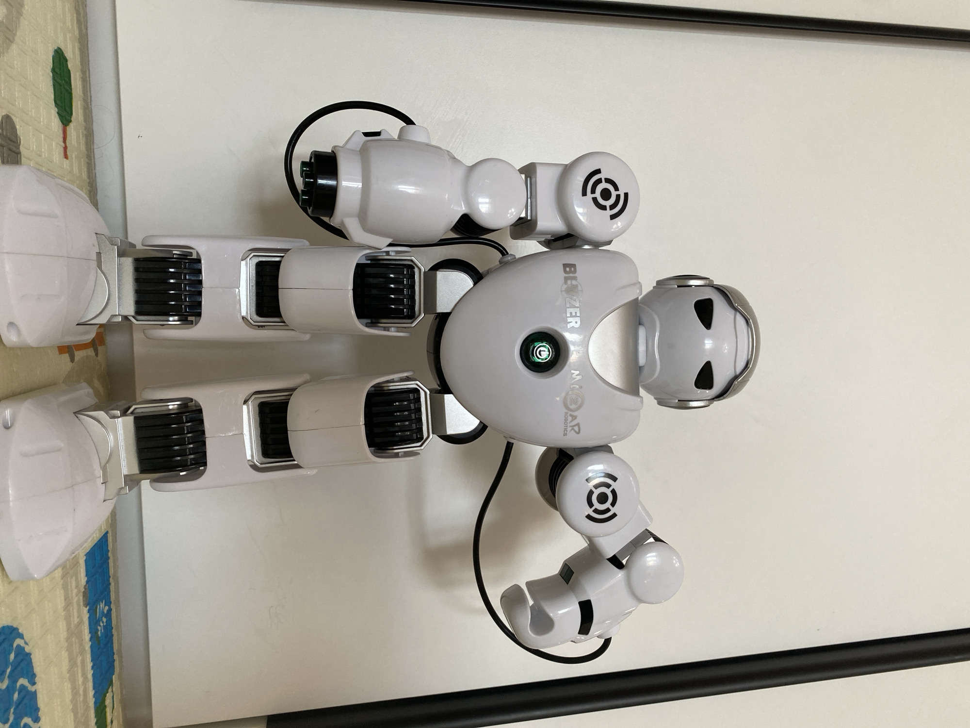 A white and gray humanoid robot stands vertically against a plain white wall. The robot has a rounded head with two dark eye-like cutouts and a silver trim around the top. Its torso is smooth and features a green light in the center, with the word "BLAZER" printed above and "MizAR ROBOTICS" printed below. The robot's arms are articulated with multiple joints and cylindrical components. Its legs are also segmented and feature black ribbed sections. A black cable extends from the robot's torso. A black horizontal bar is visible at the top of the frame. The bottom left corner shows a portion of a patterned mat. The location in the city of Unknown, Unknown is not discernable from this image.