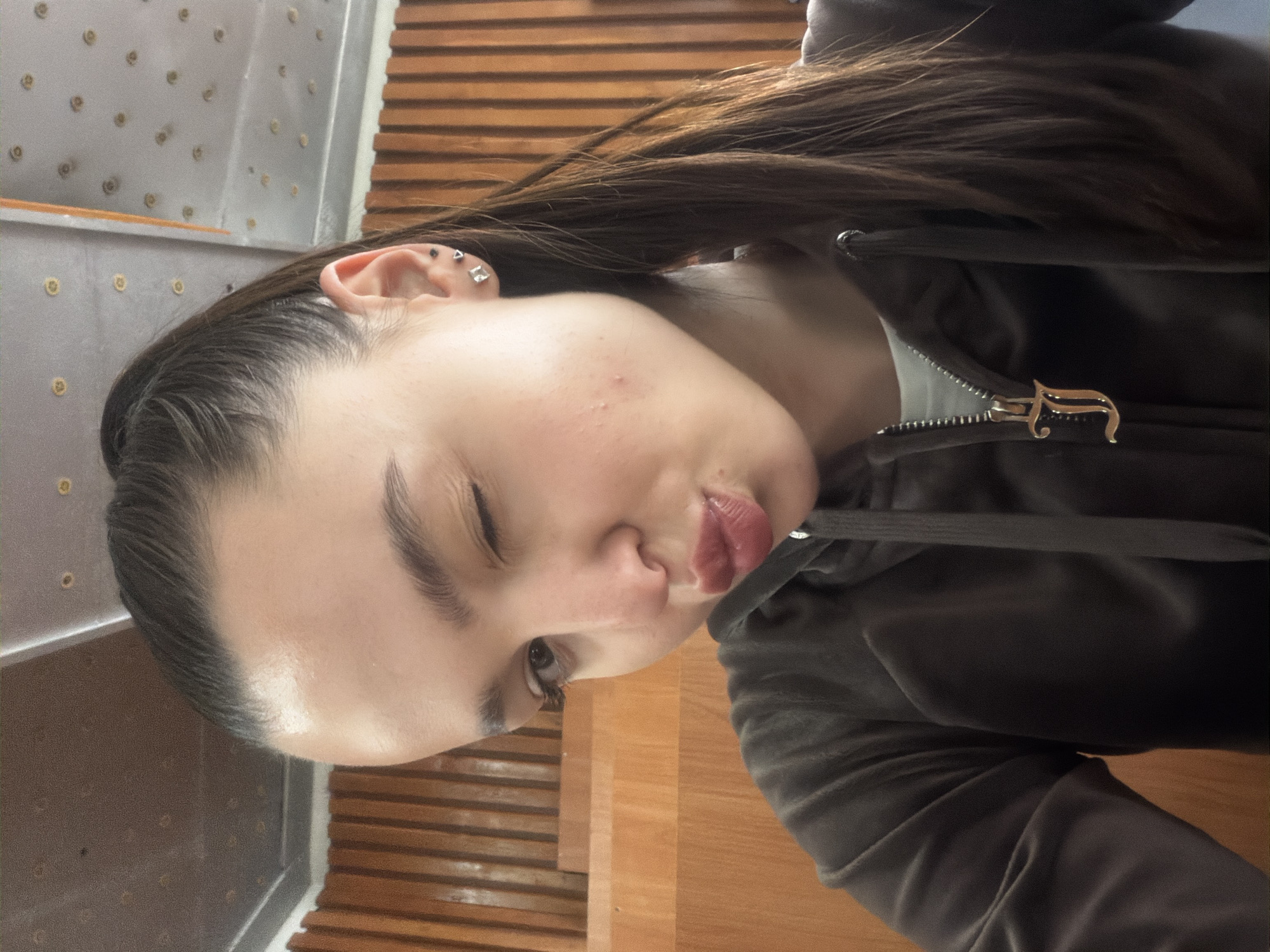 A portrait-style image, rotated ninety degrees clockwise, captures a young person with long, dark brown hair against an indoor background. The background features two distinct sections: one side displays a light-colored, textured wall or paneling adorned with small, evenly spaced circular metallic embellishments, while the other side shows horizontal wooden slats, possibly part of a wall or floor.
The subject is wearing a dark-colored (likely black or dark gray) hooded jacket or top, made of a soft, possibly velvet or velour fabric. A prominent gold-colored zipper pull, intricately shaped like the letter 'J', is visible on the jacket's front zipper.
The person is looking towards the viewer with a playful and expressive demeanor. Their lips are pursed in a 'pout' or 'kissy face,' and their right eye (from the viewer's perspective) is closed or heavily squinted, while the left eye is open. Several small, metallic earrings are visible on the left ear, including a triangular stud and a square stud. The hair is parted and slicked back from the forehead, framing the face. The skin on the forehead and temple areas shows some minor texture and small blemishes. The lighting is soft and consistent, indicative of an indoor setting. The image focuses on the person's upper body and facial expression, with their clothing and the immediate surroundings providing context.