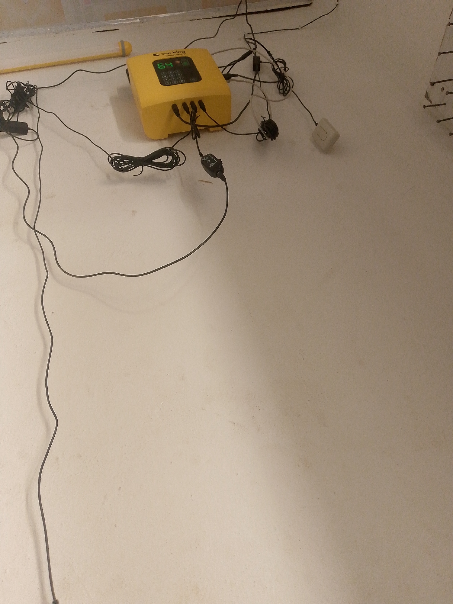 A yellow electronic device with a digital display is situated on a white floor. The device has several cables connected to it. To the left of the device, a yellow rod with a circular yellow component at one end is positioned on the floor, also connected to some of the cables. The environment appears to be an indoor space with a plain, light-colored wall in the background. The overall scene suggests a setup related to electronics or potentially environmental monitoring, but there is no contextual information to place it specifically in Jauro Yinu, Nigeria.