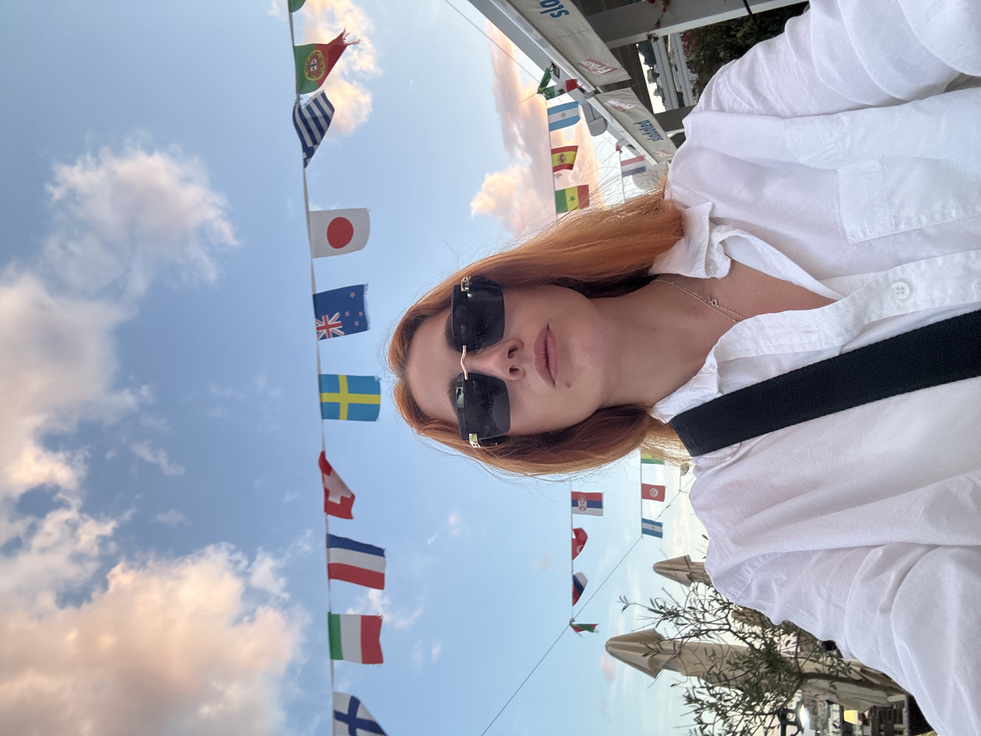 A vertical, medium shot shows a woman in sunglasses and a white shirt standing outside with a backdrop of international flags hanging on a string. The sky is partially cloudy and a light blue color. The woman has red hair and is facing the camera, with her head slightly tilted. She is wearing a gold necklace. The flags include those of Portugal, Greece, Japan, New Zealand, Sweden, Switzerland, France, Italy, and Finland. In the lower right corner, there are trees and umbrellas. The location is Budva, Montenegro.