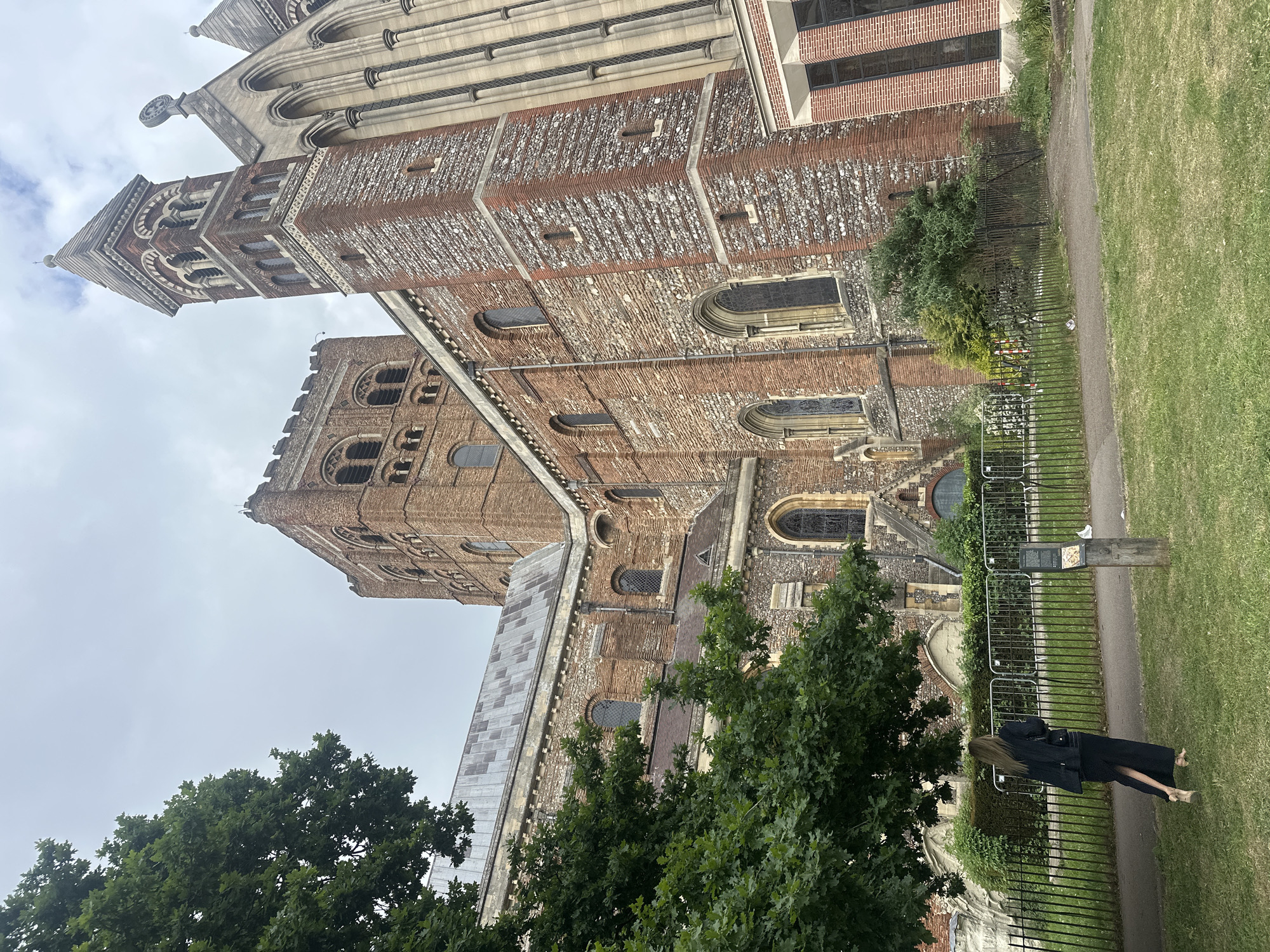 The image shows the Cathedral and Abbey Church of Saint Alban in St Albans, United Kingdom, viewed from ground level. The church is a mix of brick and stone construction, with a tall tower visible. A woman stands along a path separated from the church grounds by a black metal fence. The path is adjacent to a grassy area. Trees are present, partially obscuring the lower portions of the church. The sky is overcast.