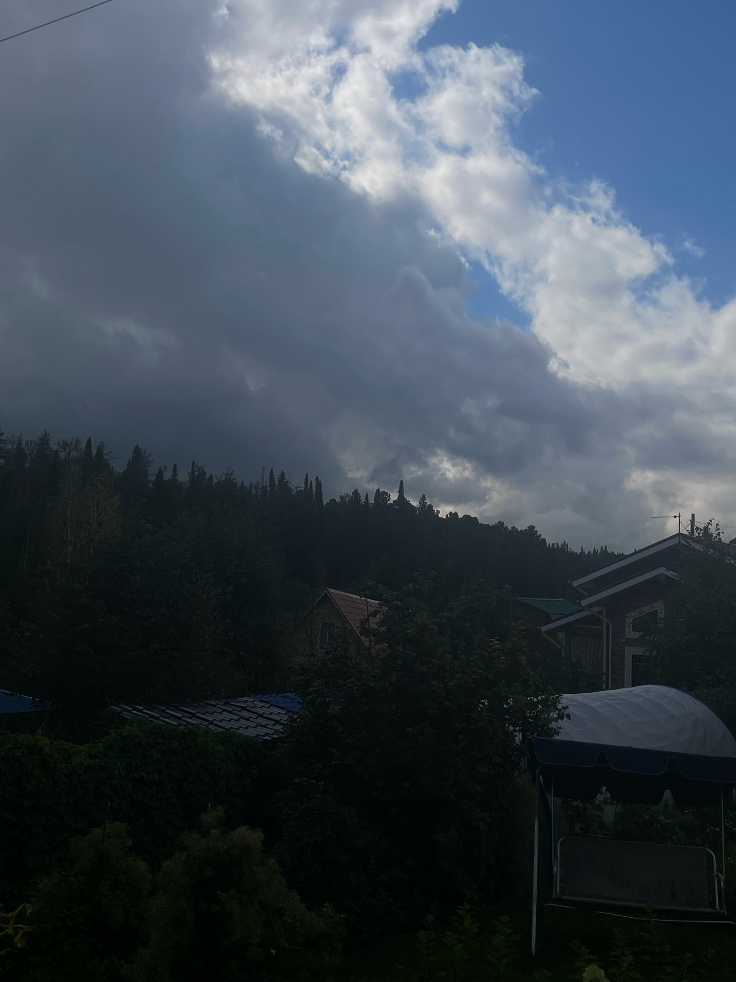 The image depicts a view of a hillside with dense trees and several structures in Bogashevo, Russia. The sky is filled with dramatic clouds, with patches of blue visible. In the foreground, a dark swing set with a blue canopy is positioned. Behind the swing set, obscured by foliage, are the roofs of two buildings. One building has a dark tiled roof, and another, further up the hill, has a red-tiled roof. The hillside is covered in dark green trees, with the silhouettes of taller trees visible against the sky on the ridge. The overall atmosphere is somber due to the heavy clouds and shadows.