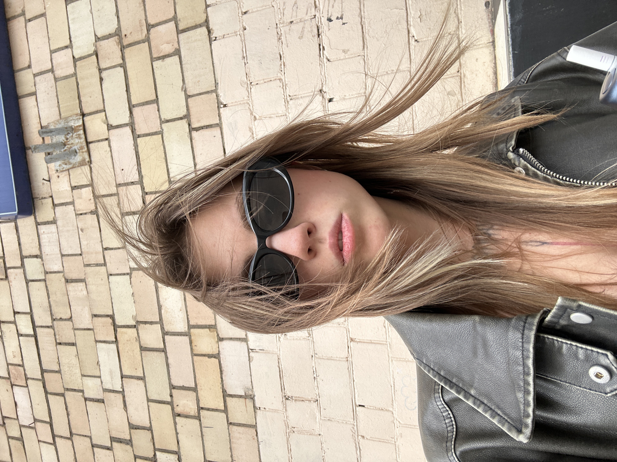 A young woman wearing sunglasses and a leather jacket stands against a light-colored brick wall. Her hair is blowing in the wind. The scene appears to be outdoors in a city environment. The brick wall is textured and shows signs of age, with some discoloration and minor debris. A metal fixture is attached to the wall above and to the right of the woman's head, and a portion of a blue sign is visible at the very top of the image. The woman is looking directly at the camera. The overall impression is a candid street-style portrait.