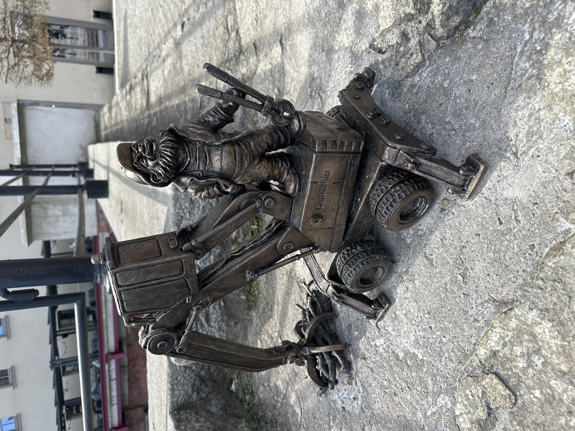 A bronze dwarf statue of a construction worker operating an excavator is positioned on a concrete surface. The dwarf is seated on the vehicle, holding tools. The excavator is detailed, with wheels, arms, and a cab. The concrete surface has natural variations in texture. In the background, there is a multi-story building with visible windows and a railing. The statue is located in Wrocław, Poland, known for its dwarf statues.