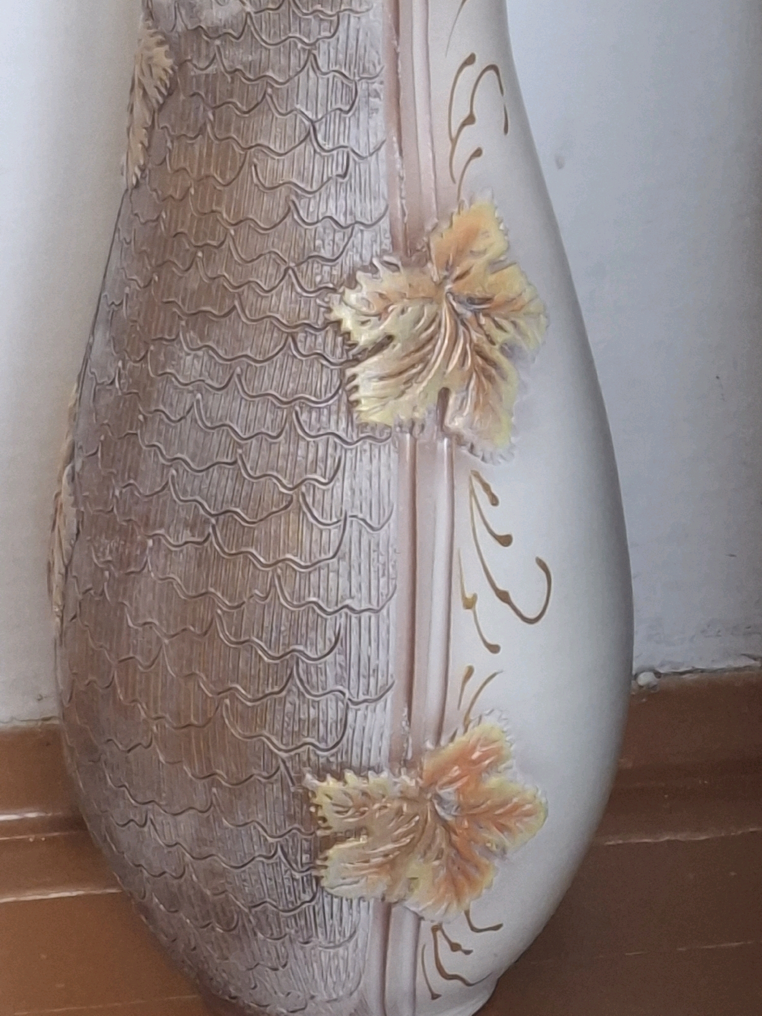 The image shows a close-up view of a decorative vase. The vase has a split design, with one side featuring a textured, scale-like pattern in shades of brown and beige. The other side of the vase is smoother and lighter in color, with embossed floral or leaf motifs in yellow and orange, outlined in gold. The vase is leaning against a white wall, with a wooden floor visible at the bottom. The lighting suggests an indoor setting. There are no people or animals present. The location in Almaty, Kazakhstan is not specified by the image.