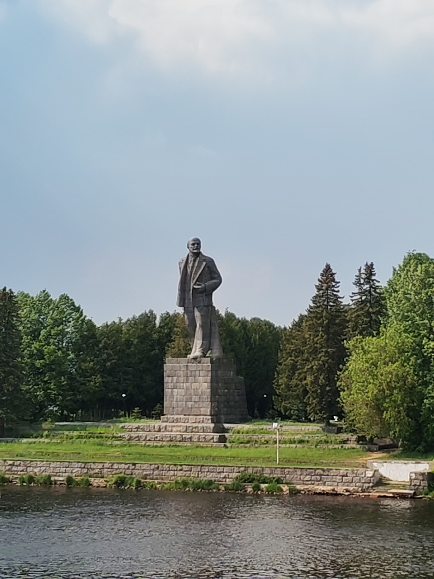 The image shows a statue of Vladimir Lenin standing on a tall, square pedestal. The statue and pedestal are located on a grassy area near a body of water, possibly a river or lake. Trees surround the statue and grassy area. The sky is light blue with some clouds.