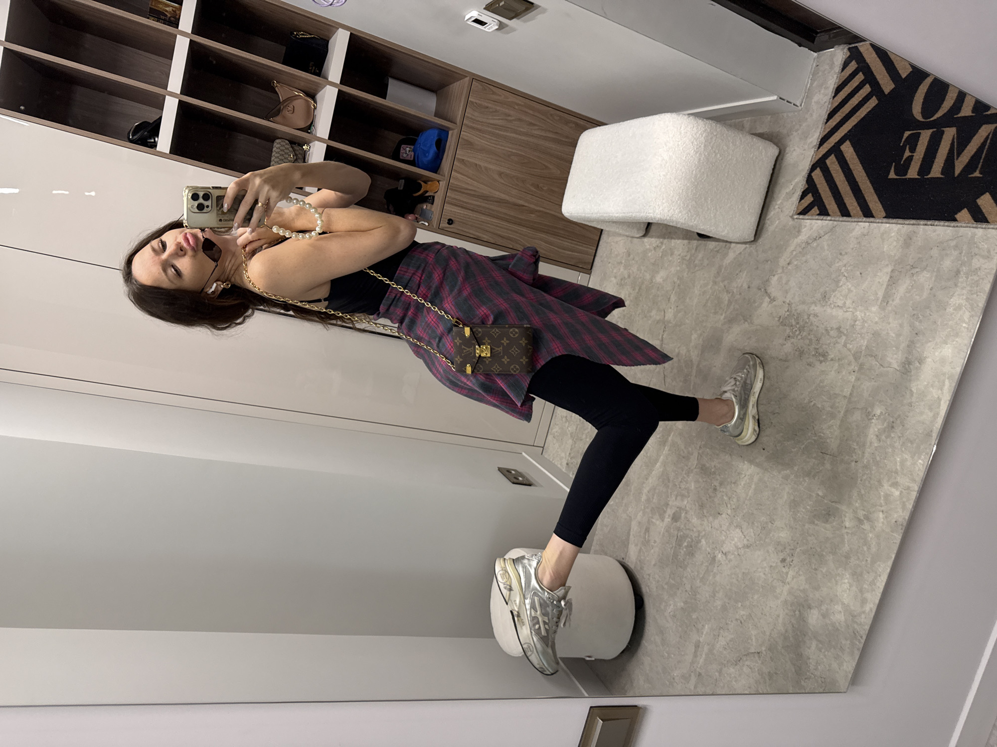 In a mirrored room, a young woman takes a selfie, holding her phone and striking a pose. She is wearing a black tank top, a pink and grey plaid shirt tied around her waist, black leggings, and silver sneakers. A small Louis Vuitton purse with a gold chain hangs across her body. She adorns herself with pearl necklaces and earrings. The background reveals shelves filled with bags and accessories, a white ottoman, and a doormat with the word "WELCOME" visible. The marble-tiled floor reflects the scene. The lighting suggests it is daytime. The woman appears playful and confident. The location is Khimki, Russia.