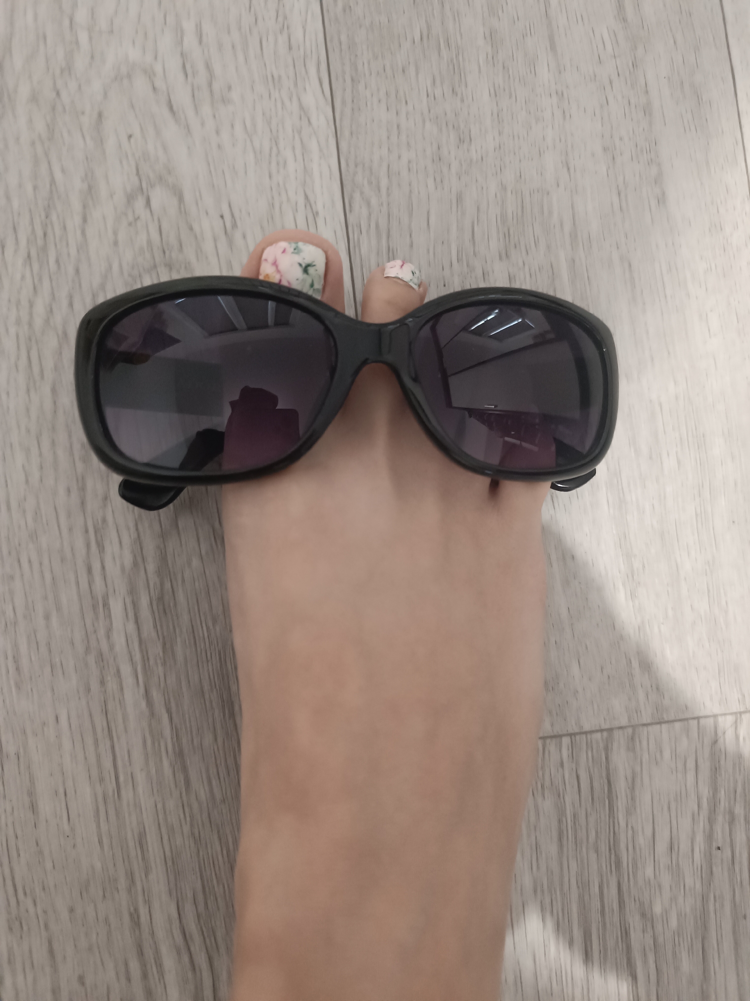 A person's foot, with painted toenails, is positioned on a light-colored, wood-plank floor. A pair of black-rimmed sunglasses rests on top of the foot, covering where eyes would normally be. The sunglasses are slightly angled, and reflections of an indoor ceiling light are visible on the lenses. The scene is simple and lacks any discernible urban or city elements.