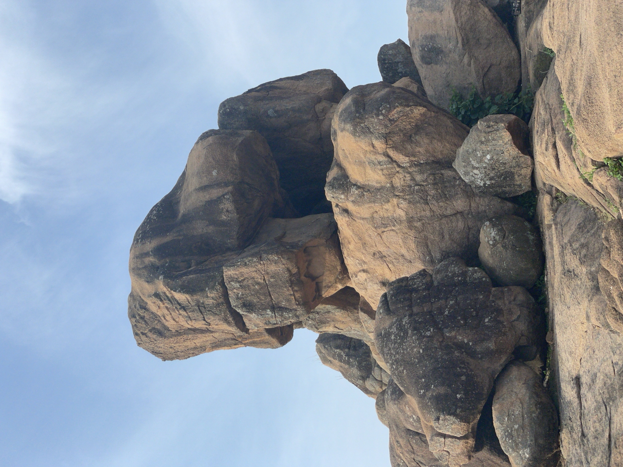 The image shows a rock formation against a blue sky. The rocks are various shades of brown and grey, with some green vegetation growing between them. The photograph appears to be taken from a low angle, looking upwards at the rocks. The location is Barikin Gidar Kanat, Nigeria.