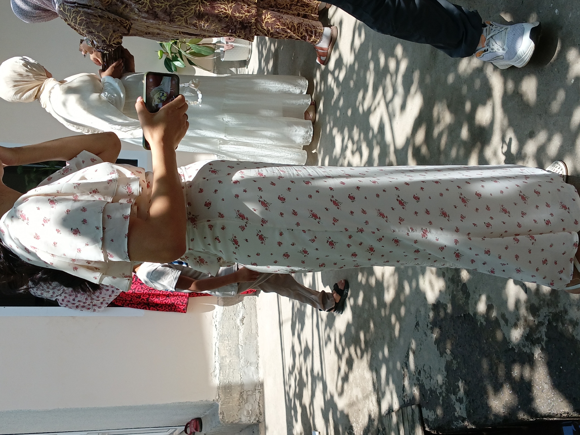 The image captures a group of people outdoors in what appears to be a city environment, possibly Osh, Kyrgyzstan, based on the hint. The setting is illuminated by sunlight, casting distinct shadows on the concrete ground, suggesting it is daytime.
In the foreground, a woman in a white floral print dress is prominently featured, her side facing the camera. She is holding a smartphone in her right hand, which is angled to display a picture on its screen. Her left hand is raised to her face, obscuring part of her features.
Behind her, another person, possibly a child, is visible wearing a white t-shirt and brown pants, with black sandals on their feet. Further back, a woman in a flowing white garment and a headscarf is seen holding a woven basket. To the right, another individual is wearing a patterned brown garment and sandals, and a third person is wearing dark pants and white athletic shoes.
The background shows a white building with some textured concrete elements and a doorway. A potted plant is visible to the right of the woman with the basket. The overall scene suggests a casual gathering or event taking place outdoors.