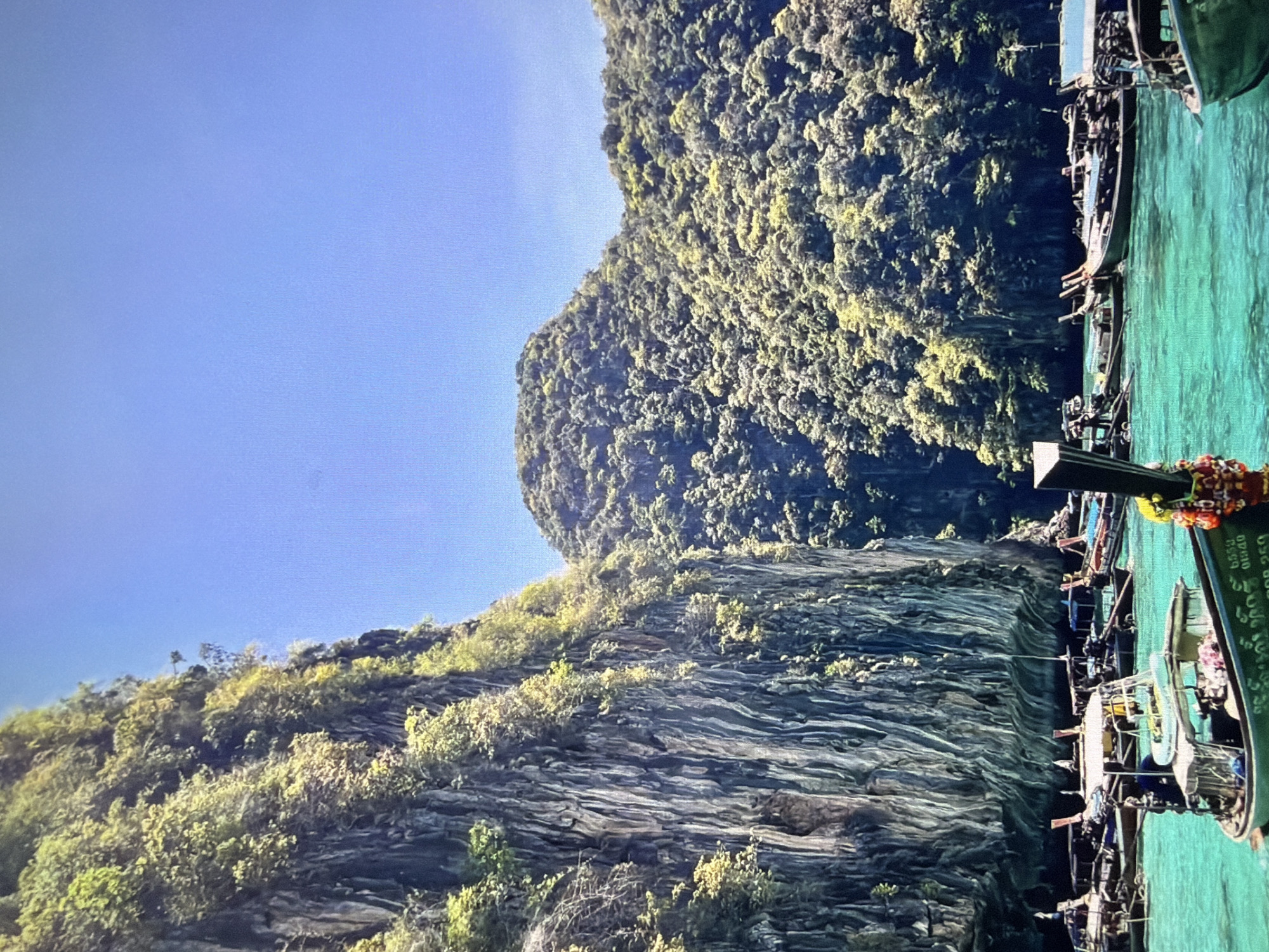 This media file presents a scenic tropical seascape, originally captured and displayed on its side. When viewed correctly, the image shows several traditional longtail boats moored on clear, turquoise-emerald water. Towering, lush green karstic limestone cliffs and islands, densely covered in vegetation, rise dramatically from the water on the left side of the frame, extending towards the center and background. The sky above is a bright, unblemished blue, indicating clear, sunny weather.
In the foreground, several longtail boats are visible along the right edge of the image. The closest boat is a deep green, adorned with colorful garlands or cloths in red, orange, and yellow hues near its bow. On the side of this boat, white text is visible, including the numbers "6333" and "9940", and below that, "250". Some indistinct Thai script also appears. Other longtail boats are partially visible behind the lead boat, suggesting a group or popular mooring area.
The scene depicts a calm, sunny day, likely in a well-known Southeast Asian coastal region, such as Thailand, due to the distinctive longtail boats and limestone formations. The activity appears to be tourist-related transport or leisure, with boats awaiting passengers or anchored for enjoyment of the stunning natural surroundings. The overall impression is one of natural beauty and tranquility.