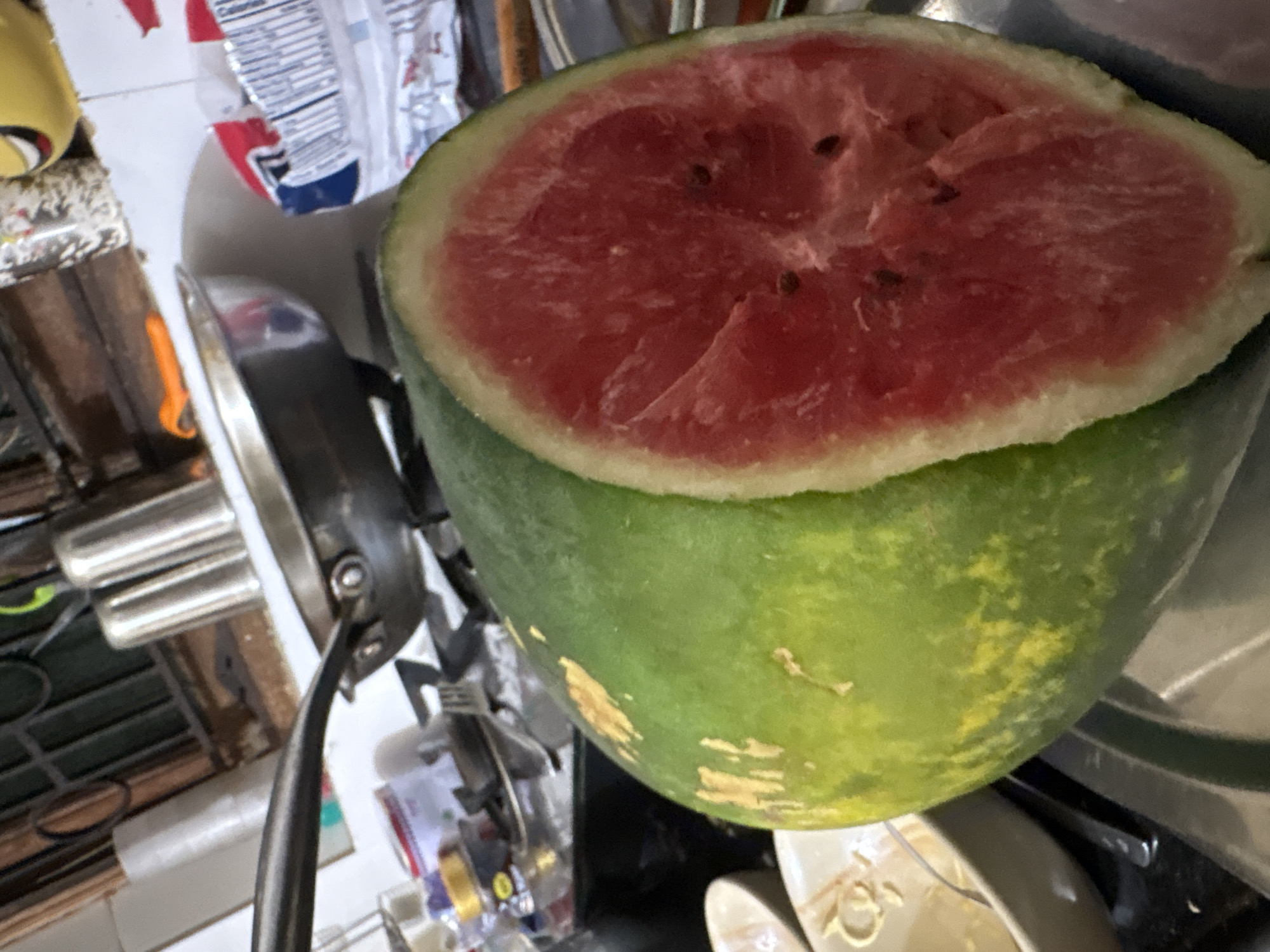 The image depicts a kitchen counter in Ashvem, India, with a large, halved watermelon prominently displayed in the foreground. The watermelon's vibrant red flesh and dark seeds are clearly visible. Behind the fruit, the counter is cluttered with various kitchen items, including pots, pans, and utensils, some of which appear to be on a stove. A white tiled backsplash runs along the back wall. The overall impression is of a utilitarian kitchen environment.