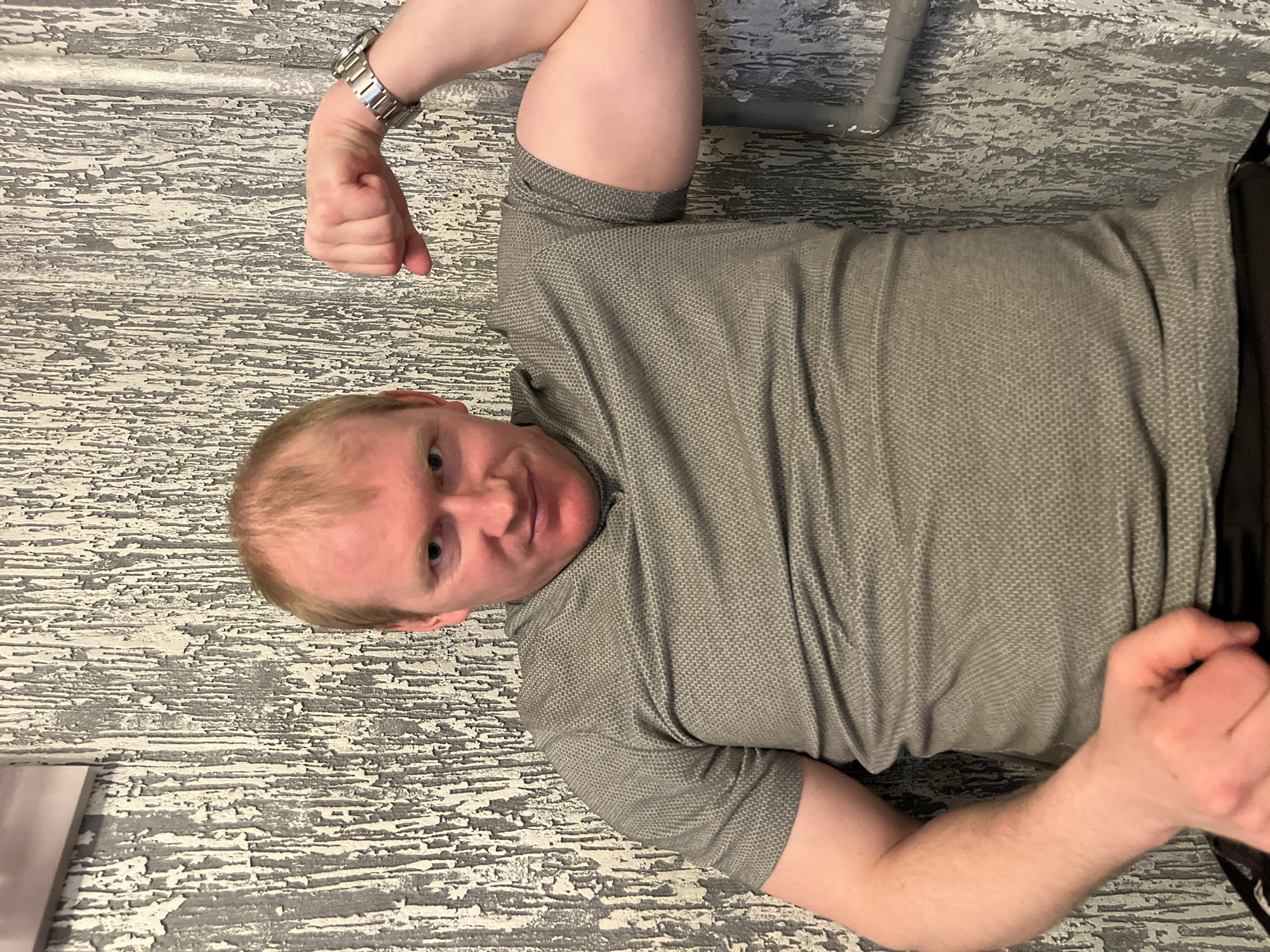 The image displays a young man lying down and posing for the camera in an indoor setting. He has light reddish-blonde hair, fair skin, and blue eyes, looking directly at the viewer with a neutral to slightly playful expression. He is wearing a grey short-sleeved polo shirt with a distinct textured pattern and black bottoms. A silver-toned watch with a metal link band is visible on his left wrist. His left arm is bent, and he appears to be flexing his bicep with a fisted hand. His right arm is also bent, with his hand resting near his waist.
The background features a highly textured surface, possibly a wall or floor, with a distressed grey and white paint finish that gives the appearance of peeling or cracked paint. In the upper right portion of the image, a grey pipe runs horizontally across this textured background. A corner of a light-colored object, potentially a piece of paper, can be seen in the lower left. The lighting is consistent with an indoor environment, likely under artificial light, as no specific time of day or weather conditions are evident. The event captured is a casual photo opportunity. No visible text is present in the image. The location context for this media file is Chaikovsky, Russia.