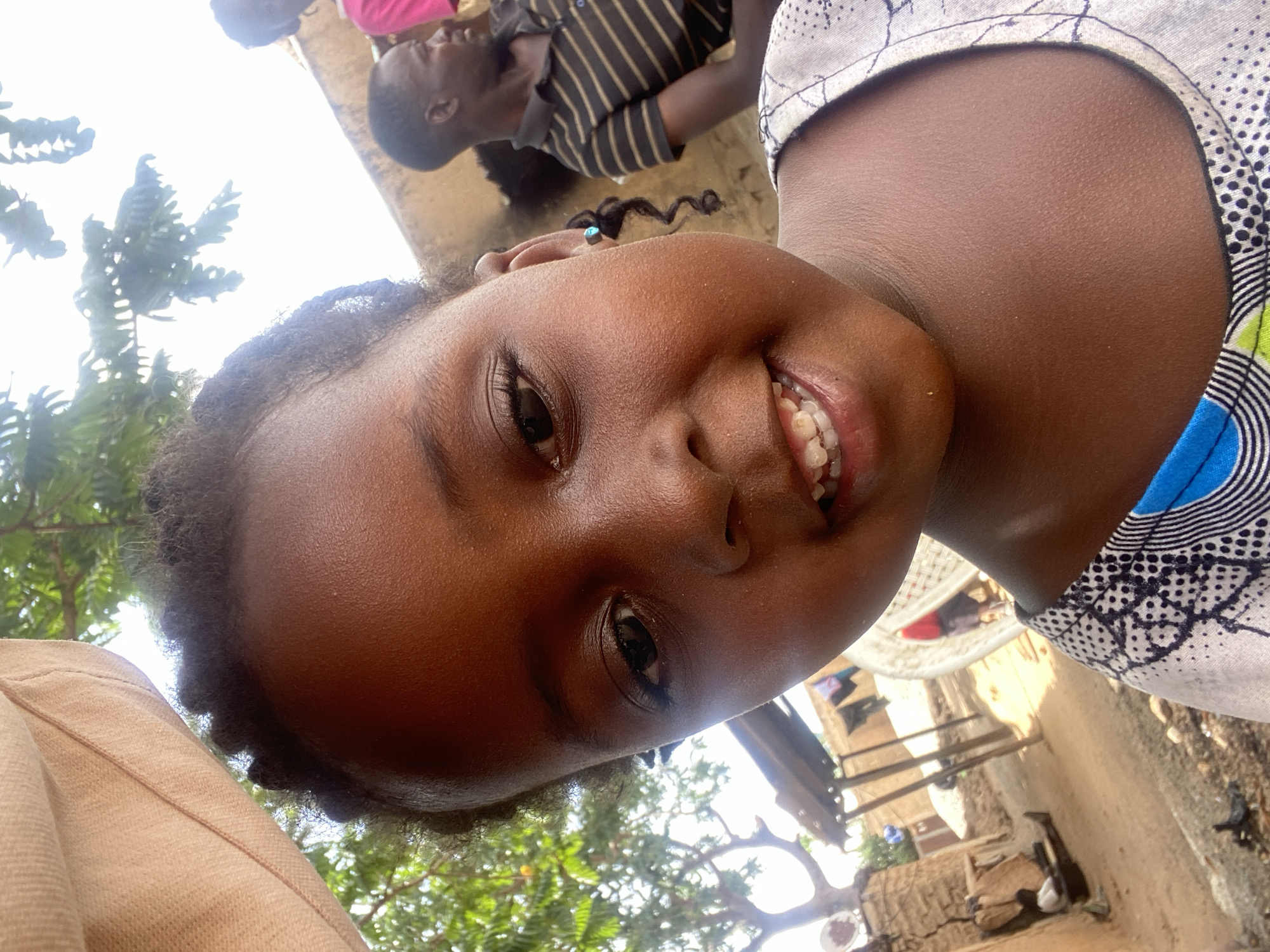The image captures a candid, joyful moment, likely taken from a low angle or as a selfie, featuring a young child with dark skin at the forefront. The child, possibly a girl, has dark, curly hair styled in small braids or twists and is looking directly at the camera with a wide, bright smile, revealing her teeth. She wears a light-colored garment with a patterned collar featuring blue, green, and black geometric and dotted designs. Her face fills a significant portion of the frame, conveying a sense of warmth and happiness.
The setting appears to be outdoors in Kuru Dungus, Nigeria, under bright daylight. In the background, another child with dark skin is visible, lying down or reclining, wearing a black and light-colored striped shirt. Further back, hints of a rustic environment can be seen, including earthy tones of soil or unpaved ground, and what appears to be simple building structures or walls made of natural materials. Green foliage from trees is visible, suggesting a natural and possibly tropical surroundings. A bright sky is also visible in parts of the background, indicating clear weather. No visible text is present in the image. The overall mood is cheerful and natural.