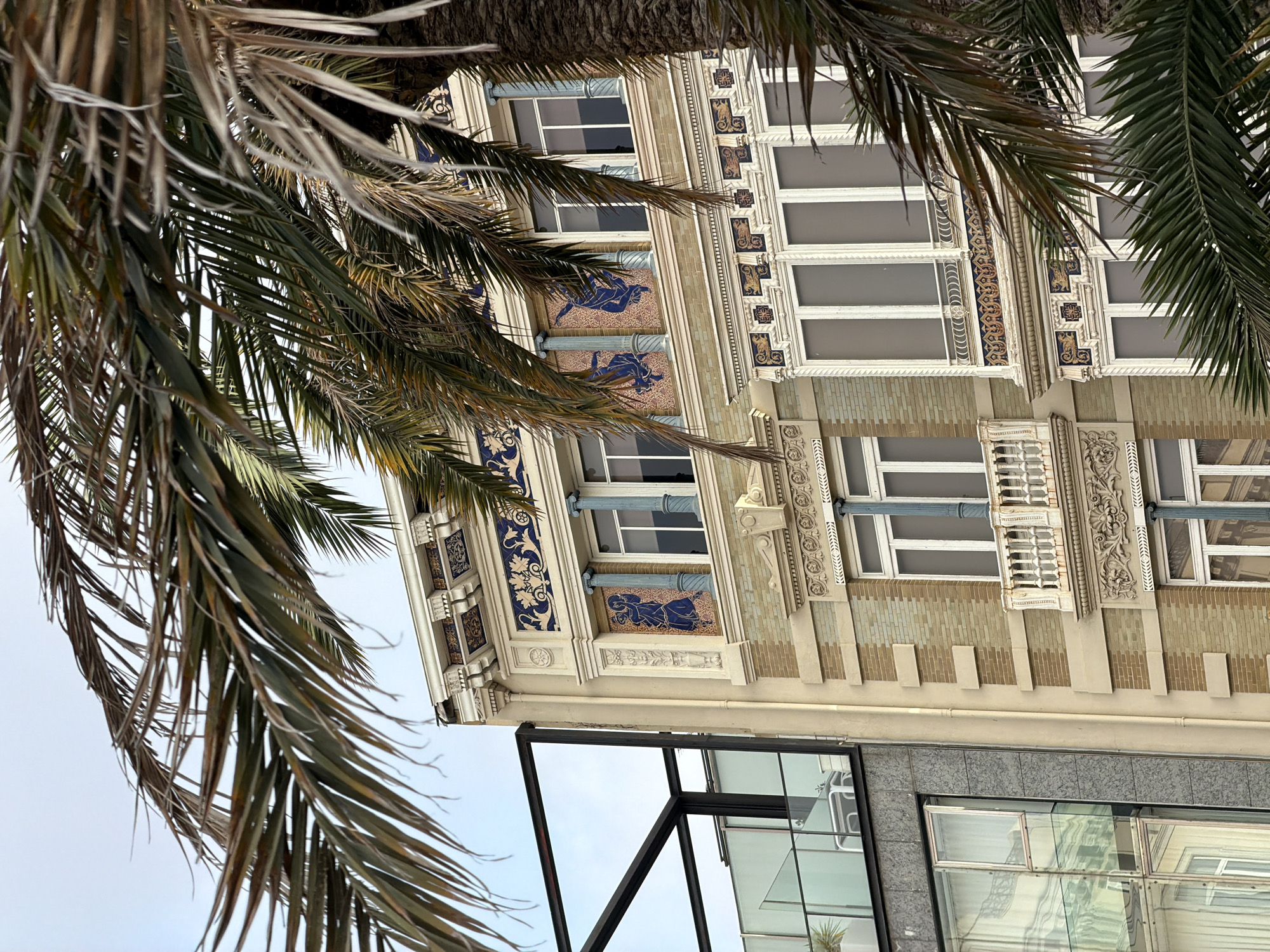 An exterior, eye-level, medium shot captures a section of an ornate building facade adjacent to a modern glass structure. The focus is on the detailed stonework, tile accents, and windows of the older building, partially obscured by the fronds of a large palm tree in the foreground.
The older building features a light beige stone base with rows of green and beige bricks. Above the brickwork, decorative moldings and elaborate tilework with blue and gold patterns are visible. Small balconies with white railings protrude from some of the windows. Two prominent windows on the upper level have blue columns and mosaic tiles depicting figures.
To the left of the older building is a modern structure with large glass panels and a black metal frame, revealing glimpses of interior spaces, including a balcony with a table and chairs. The sky above is overcast and pale. The image appears to be taken on a bright but not sunny day.