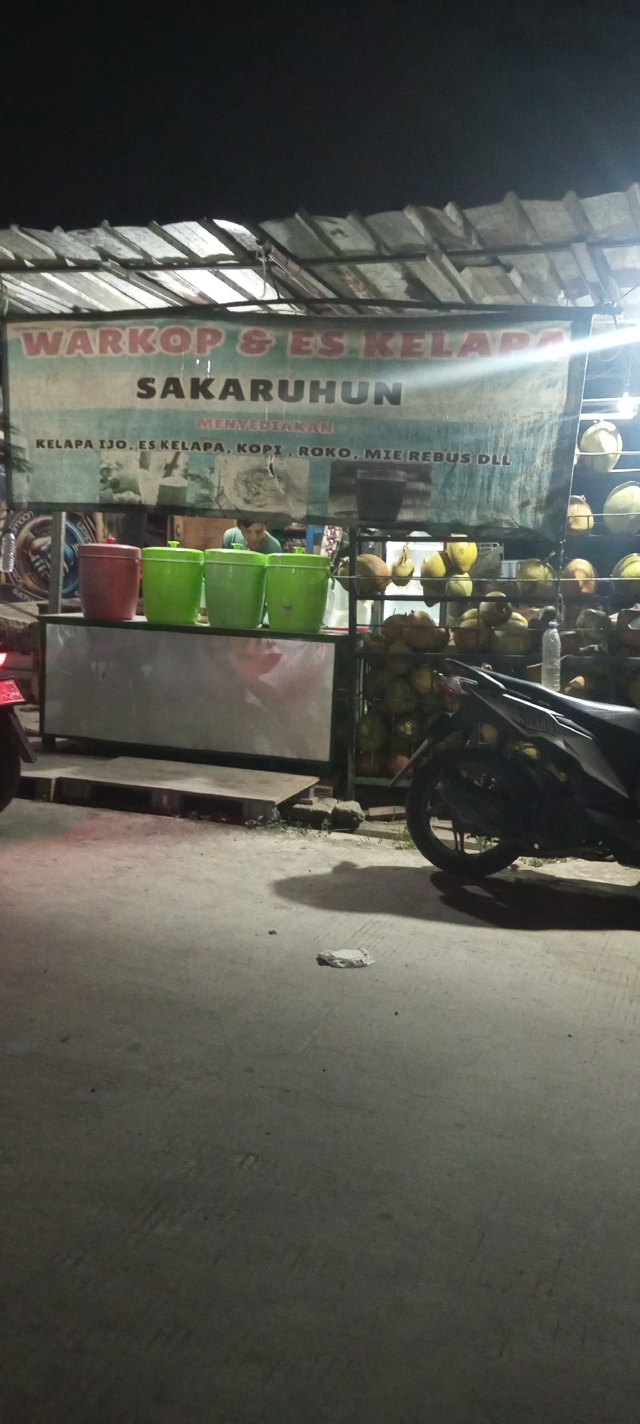 The image captures a night scene at a roadside warung in Setu, Indonesia, specializing in coffee and coconut ice. A large banner, likely for "WARKOP & ES KELAPA SAKARUHUN", is prominently displayed. The banner also lists offerings such as "KELAPA IJO, ES KELAPA, KOPI, ROKO, MIE REBUS DLL" (Green coconut, coconut ice, coffee, cigarettes, boiled noodles, etc.).
In front of the banner, a counter displays four large plastic tubs, one red and three bright green, suggesting they hold beverages or ingredients. Behind the counter, a man in a green shirt is partially visible, likely working at the establishment. A rack behind him is filled with coconuts, some whole and others split open. A motorcycle is parked to the right of the scene, its rear end and wheel clearly visible. The ground appears to be paved asphalt. The overall lighting is dim, typical of a night setting in a roadside establishment, with some artificial lights illuminating the banner and counter area.