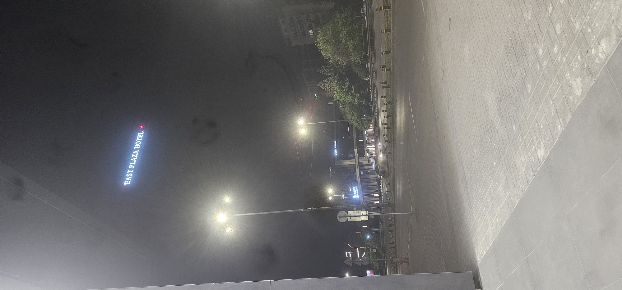 The image shows an urban scene at night in Sofia, Bulgaria. The East Plaza Hotel sign is visible in the upper left. Street lights illuminate the road and sidewalks. A building is visible in the distance, partially obscured by trees. The ground is paved with interlocking bricks and smooth tiles. There is no evidence of people in the picture. The scene appears to be a parking lot or public space near the hotel and a busy street.