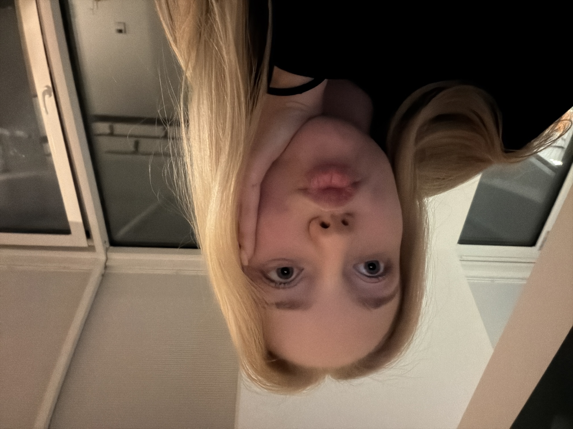 A close-up, upside-down photograph shows a fair-skinned person with light blonde hair. The person's face is tilted towards the camera with slightly pursed lips. The person has blue eyes, and one hand is positioned on the left side of their face. The person is wearing a black garment. The background includes a window frame with visible panes and a light-colored wall. The location and city are Unknown, Unknown.