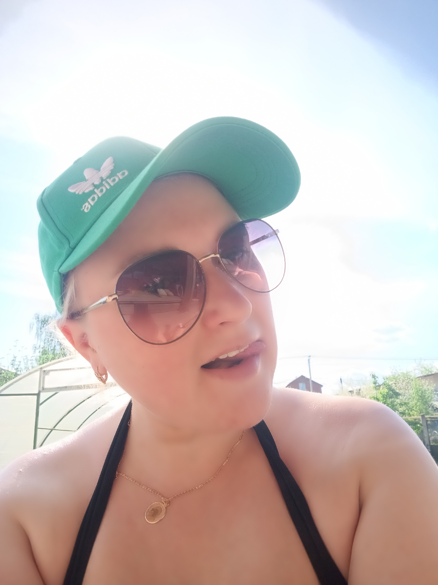 A fair-skinned woman is depicted in a selfie shot in an outdoor setting. She wears a green Adidas cap, sunglasses with gold frames, and a black bikini top. A gold necklace with a pendant is around her neck. The background reveals a greenhouse, greenery, and a house, indicative of a residential area. The location is likely Chaikovsky, Russia, based on the prompt. The lighting is bright, suggesting a sunny day. She has her tongue sticking out slightly.