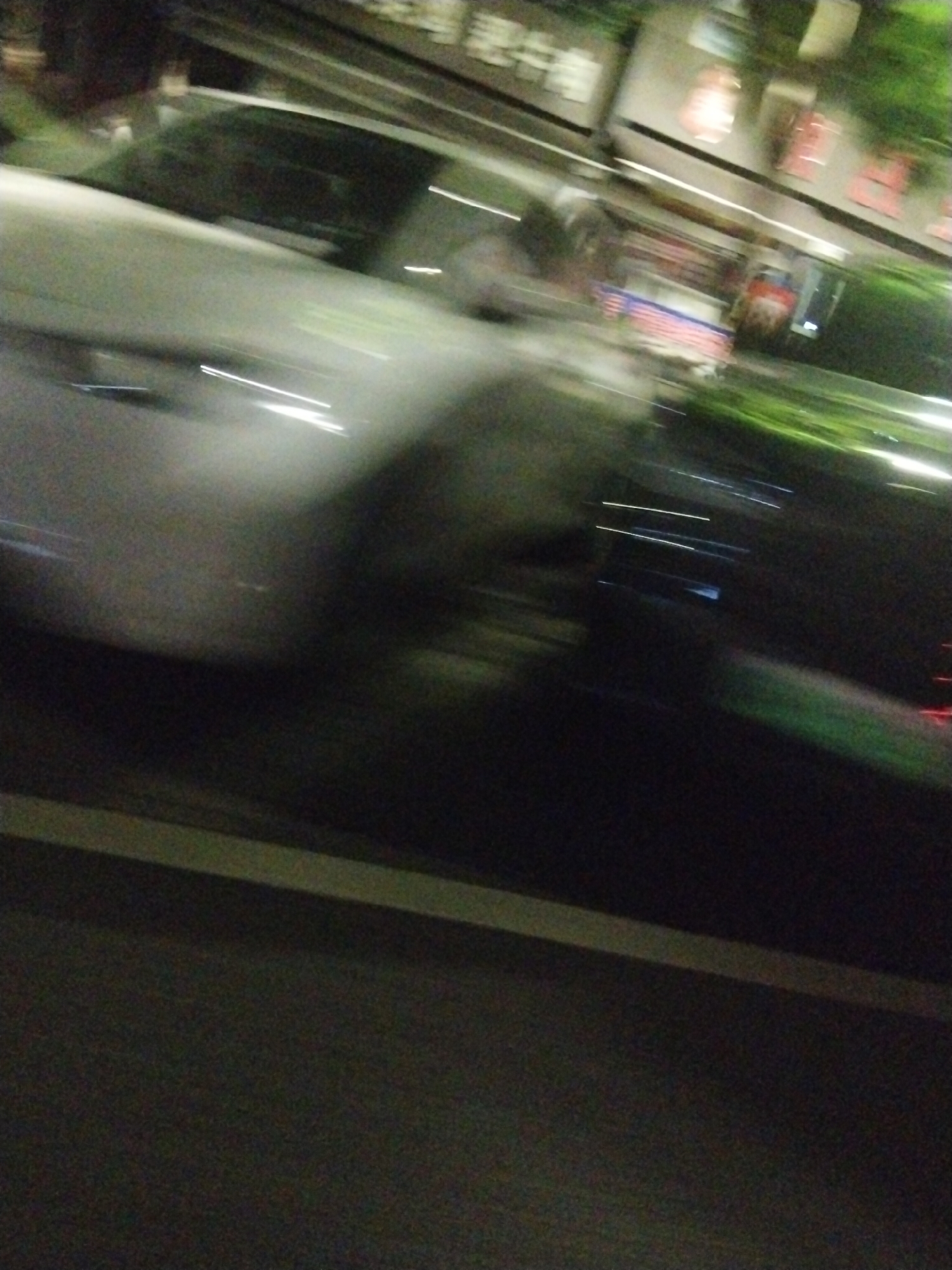 The image shows a blurry scene with a white car moving quickly along a road. The road appears to be in an urban area, possibly in the Jiulongpo District of China. A row of shops or buildings with lit windows is visible in the background. The image is motion-blurred due to the speed of the car. The road has a white line marker.