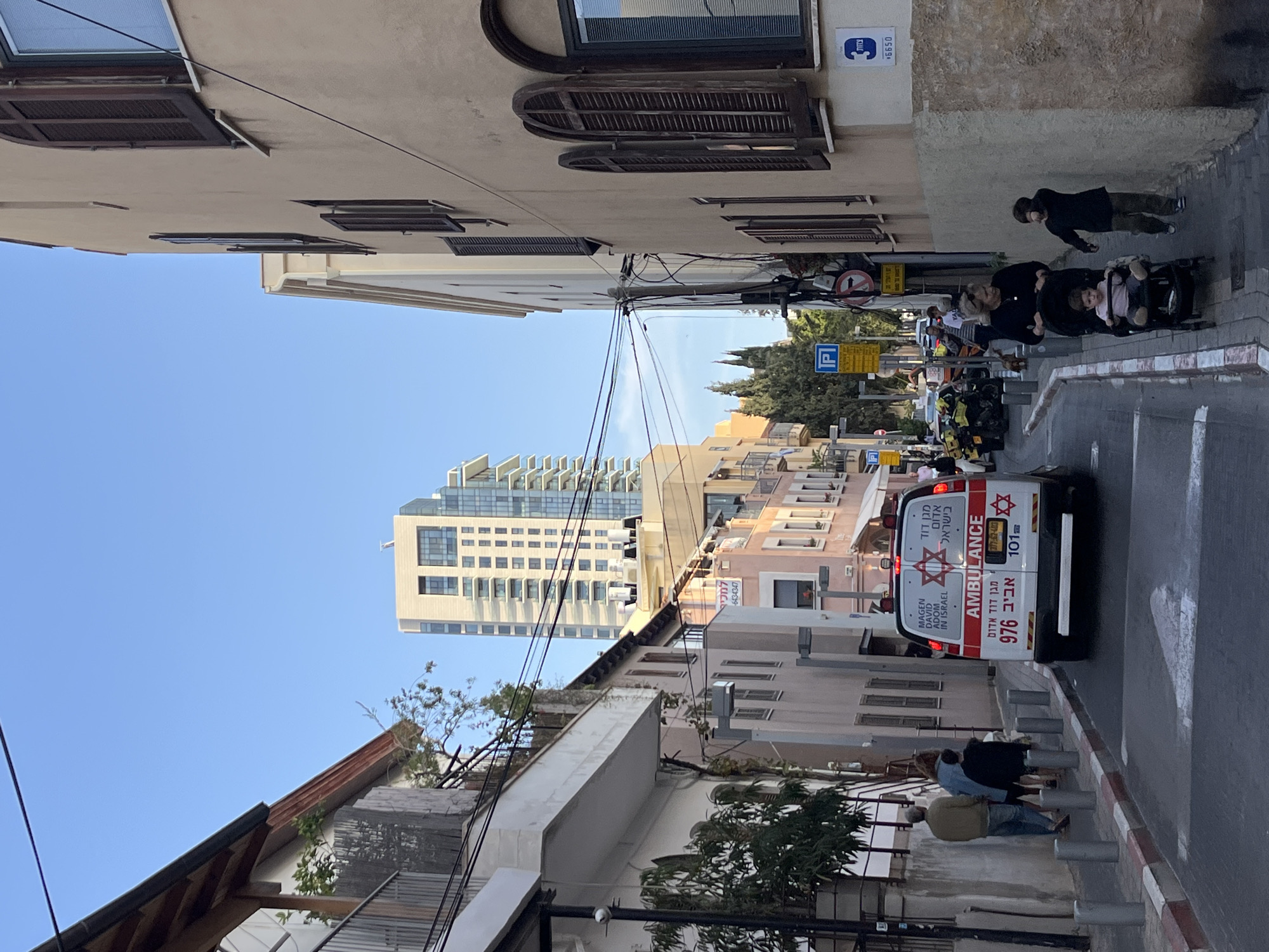 An ambulance is parked in a narrow street in Tel Aviv, Israel. People are walking on the sidewalk. In the background, a tall modern building is visible. The ambulance has the Star of David and Hebrew text on its back. There are electrical wires above the street. A parking sign and "no entry" traffic sign are visible on the side of the street. There are also signs indicating the address. The sky is blue.