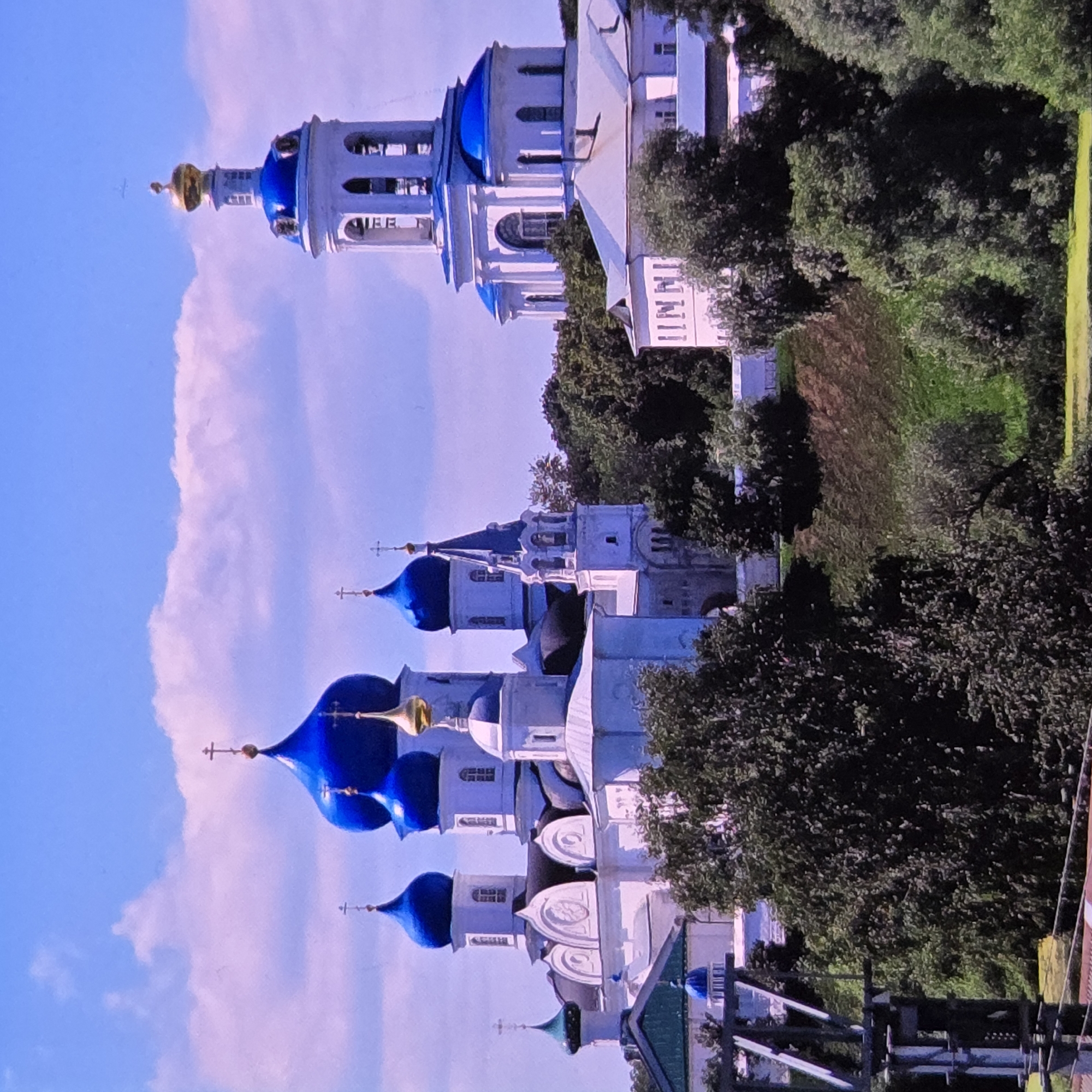 The image depicts a view of the Holy Dormition Cathedral in Vladimir, Russia. The cathedral's distinctive blue domes and white facade are prominent against a bright blue sky with scattered clouds. A tall bell tower with blue accents and a golden dome stands to the right of the main cathedral structure, which features multiple smaller blue domes topped with crosses. The cathedral complex is situated on a grassy hill, surrounded by lush green trees. In the foreground, there is a metal structure that appears to be part of a bridge or railway, and the ground is covered in grass and vegetation, with a hint of what might be water in the lower left corner. There are no people visible in the image.