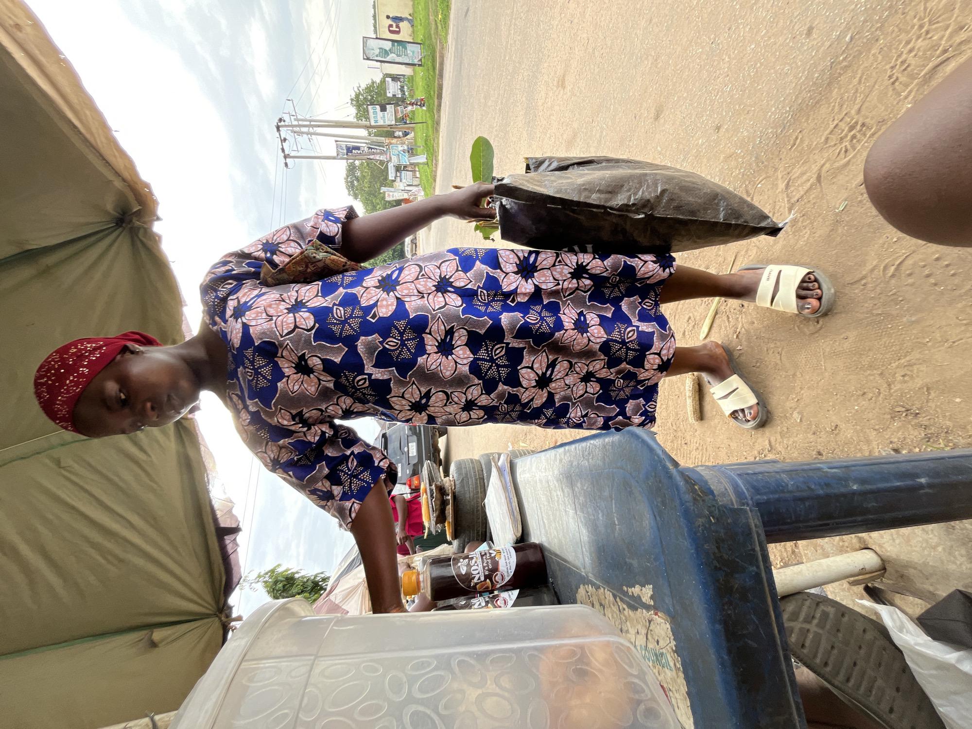 In Abuja, Nigeria, a young Black woman wearing a red headscarf and a blue and pink floral dress stands at a roadside stall under a beige awning. She is reaching for a bottle of brown liquid labeled "NOS," which appears to be a beverage or supplement. Next to the bottle is a clear plastic container holding some items, possibly food or drinks. The woman is also holding a black plastic bag containing green leaves. The stall has a blue plastic table and is situated on a dirt road with a paved road visible in the background, along with a car, utility poles, and various signs. The sky is overcast.