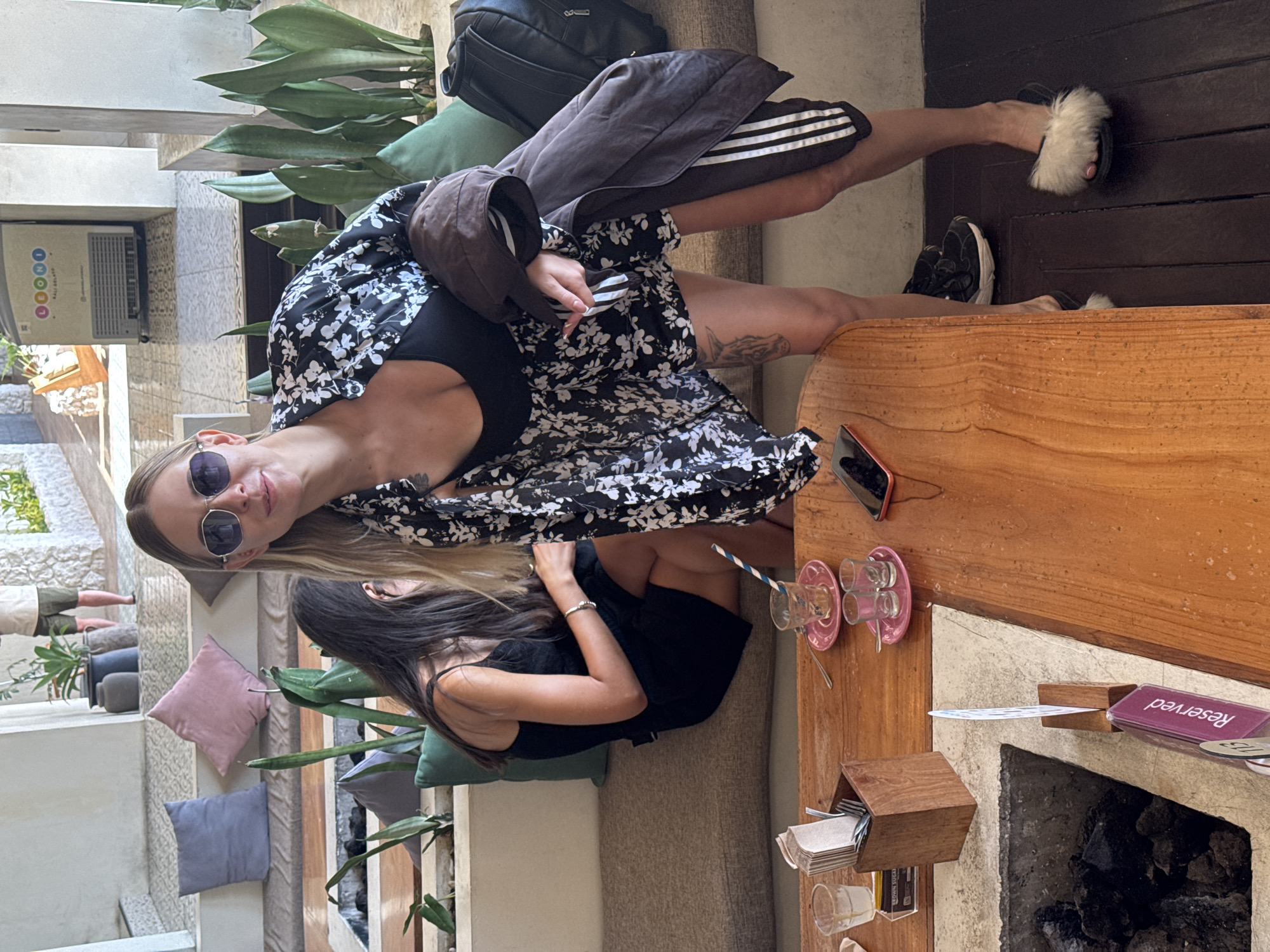 In Kintamani, Indonesia, two women are seated on a long, cushioned bench within an open-air cafe. The woman in the foreground, wearing sunglasses, a black top, and a floral print kimono-style shirt, leans forward slightly. She holds a dark brown jacket with white stripes down the arm. The second woman, seated to her left and partially obscured, wears a black sleeveless dress. In front of them is a wooden table with various items, including glasses, saucers, a napkin holder with cutlery, and a "Reserved" sign. The background reveals a modern, airy setting with white walls, potted plants, and light-colored stone accents, suggesting a stylish and contemporary establishment.
