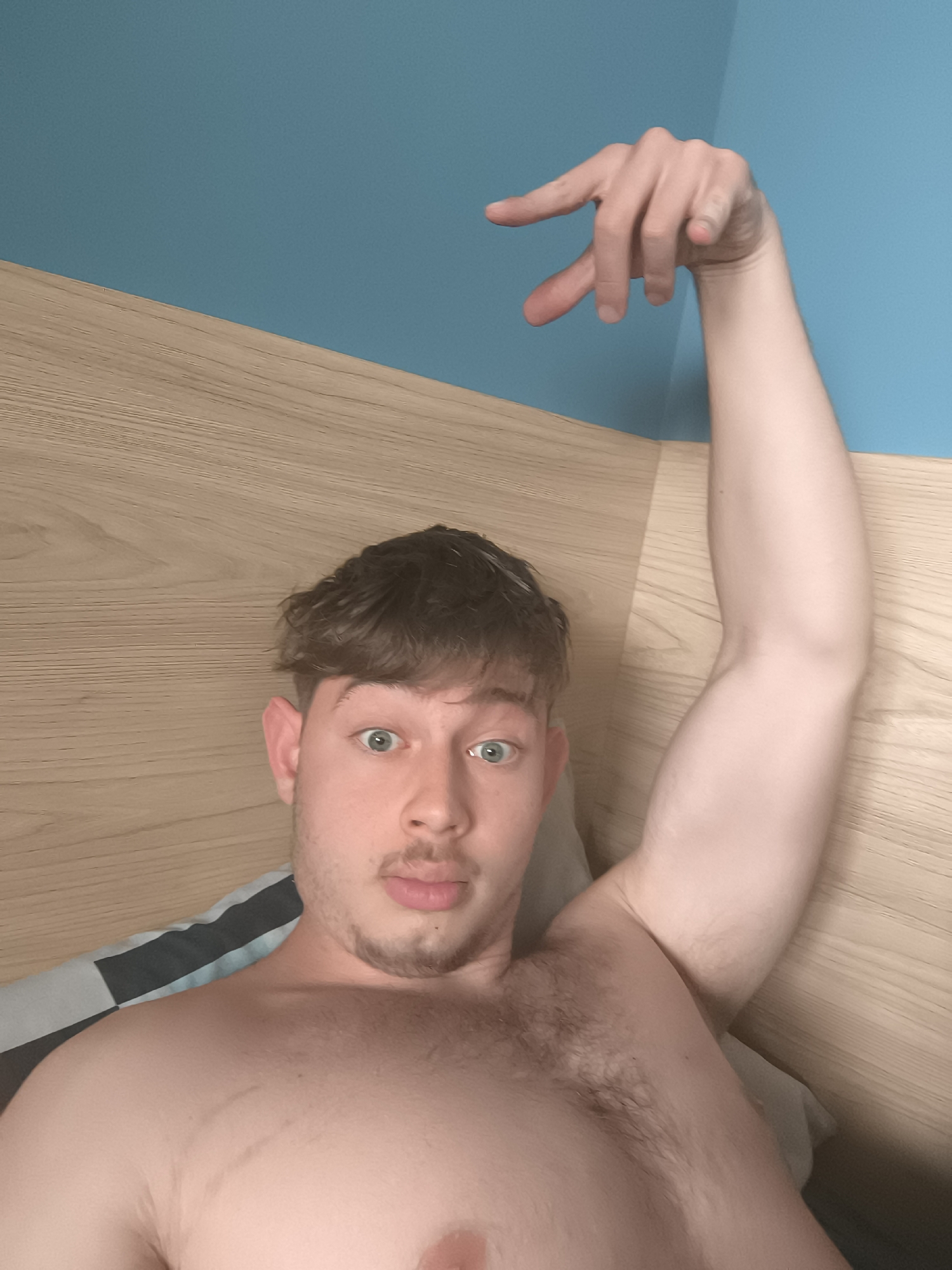 A shirtless man lies in bed with his arm raised, hand gesturing towards the blue wall. He has light brown hair and a trimmed beard. The headboard is light woodgrain. The scene appears to be indoors. There is no information to identify a specific location in Sofia, Bulgaria.