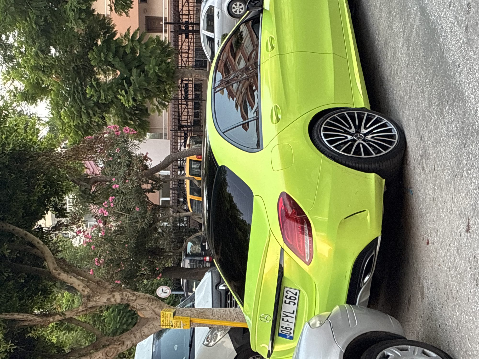 A bright lime green Mercedes-Benz sedan is parked on a paved street in Alanya, Turkey. The car has a black roof and tinted windows. The license plate, which reads "06 FYL 562," is visible. To the left of the Mercedes, a silver car is partially visible. Behind the Mercedes, several other vehicles are parked, including a yellow taxi and a white van. Trees with green foliage and some pink flowers line the street, and buildings with balconies and windows are visible in the background. A yellow sign with text is attached to a pole on the left side of the image. The scene appears to be a typical urban streetscape in a Mediterranean city.