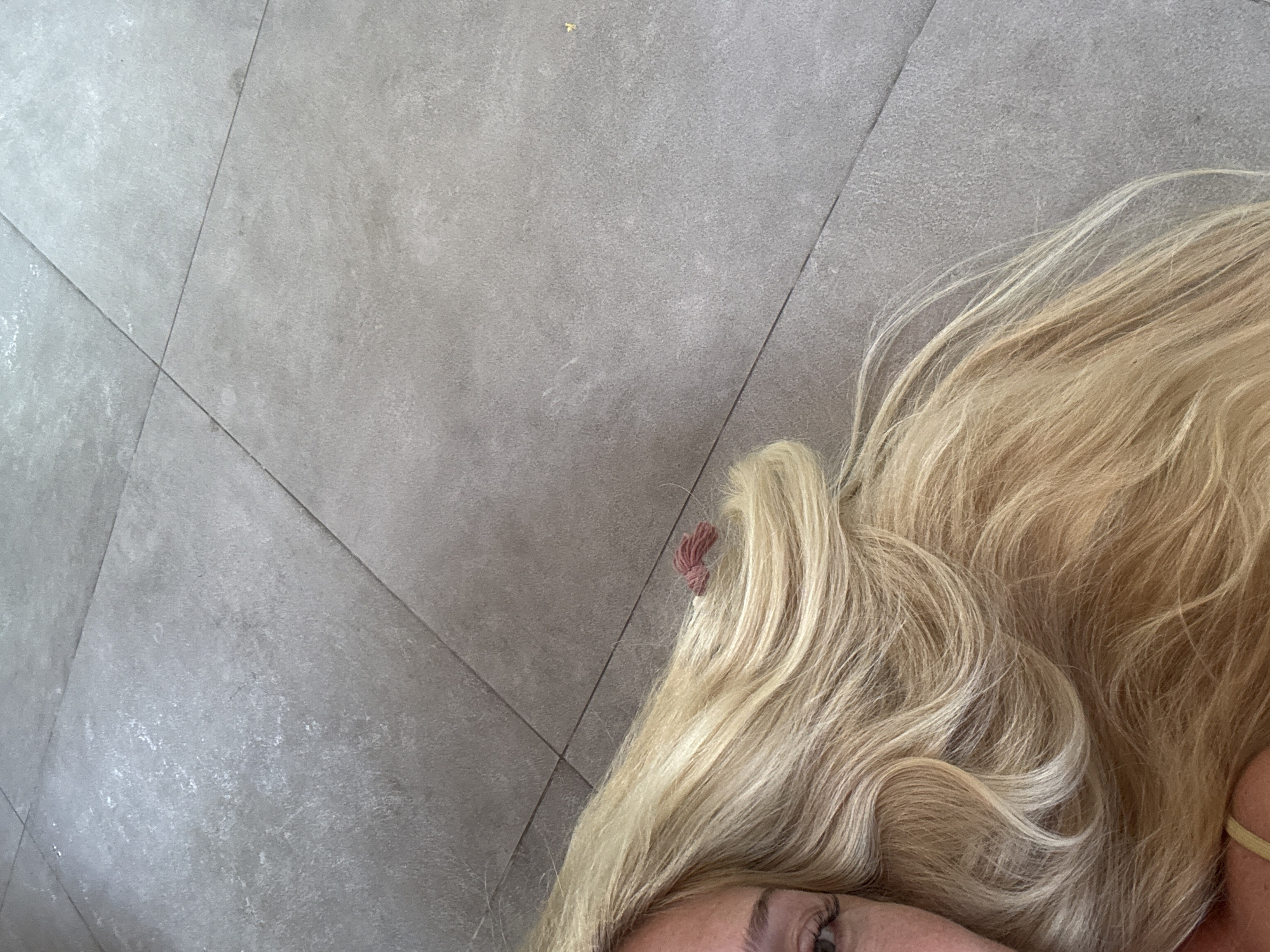 The image shows a portion of a person's face and a large mass of blonde hair lying on a grey tiled floor. A small, red hair clip is visible in the hair, near the top left of the hair mass. The lower part of an eye is visible in the bottom center of the image. The background is composed of square grey tiles, slightly textured. The location is not possible to discern.
