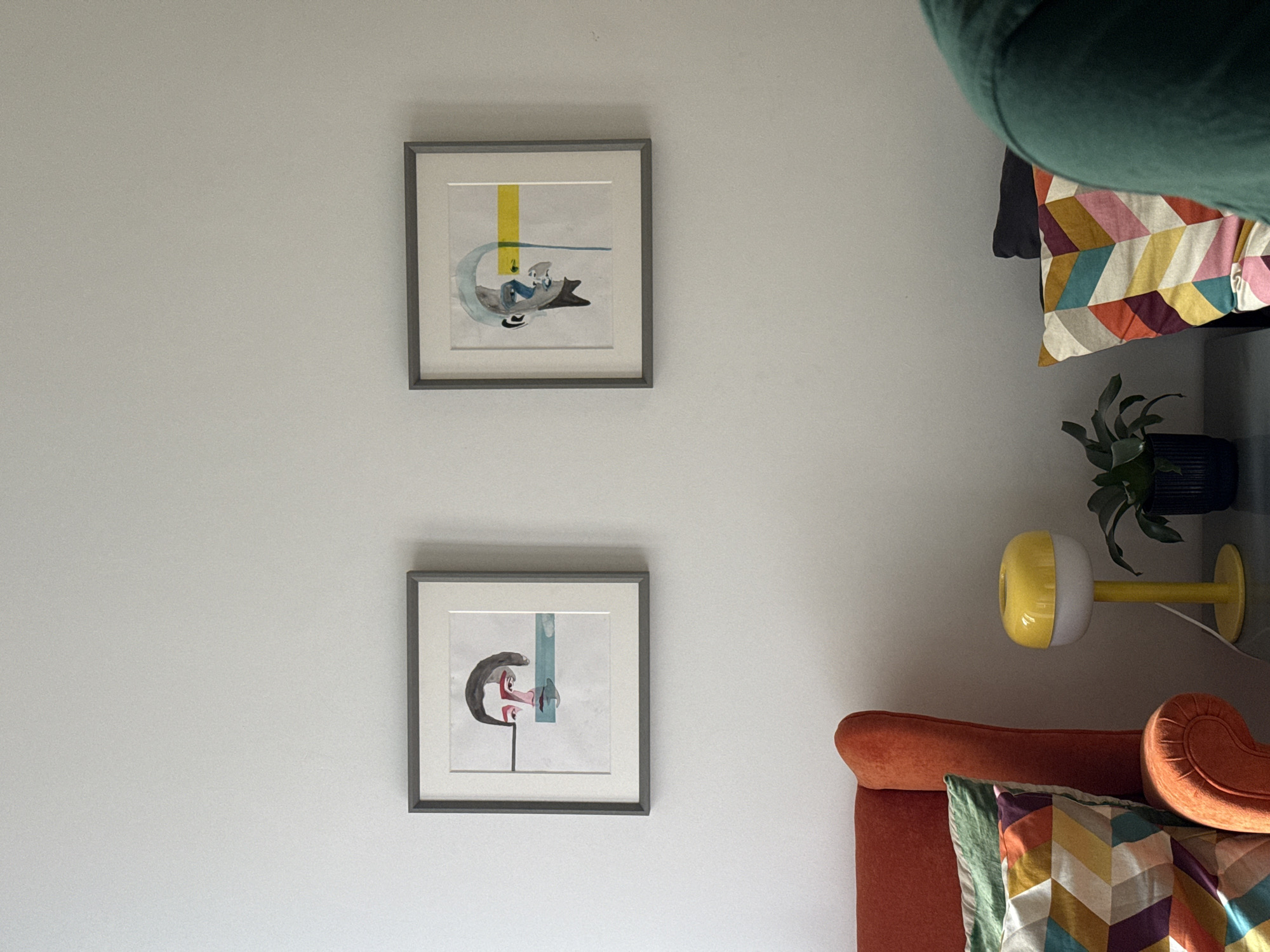 Two framed pictures hang on a white wall. The top picture features an abstract figure reclining on a crescent shape, beneath a yellow vertical stripe. The bottom picture depicts a portrait of a stylized face with a blue vertical stripe in the background. To the right of the pictures are elements of a room including a green sofa pillow, a house plant on a shelf, a lamp, and an orange armchair. Location: Unknown, Unknown.